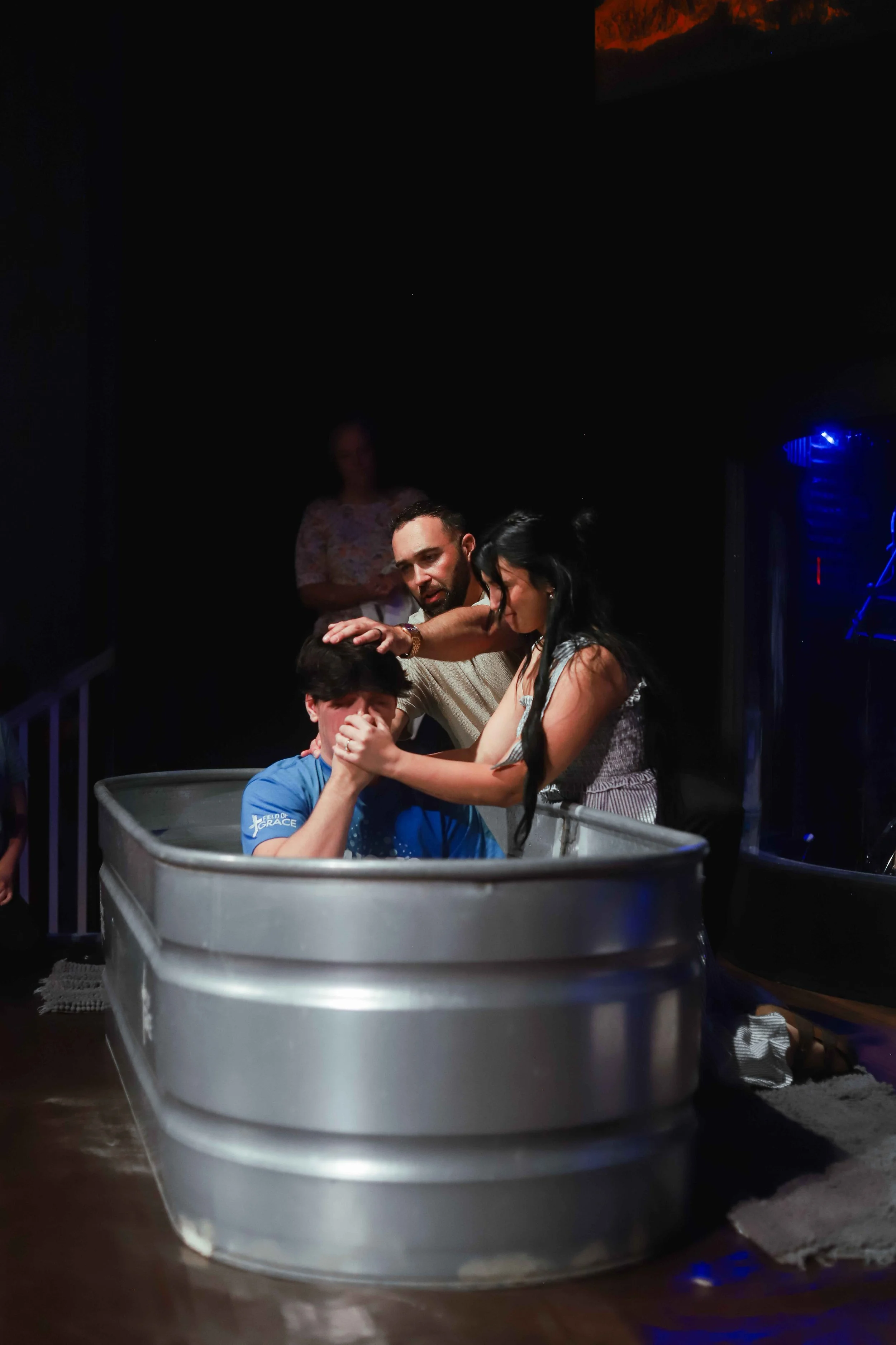 A boy is baptized in a silver metal baptism pool, with two adults helping and supporting him during the prayer and immersion.