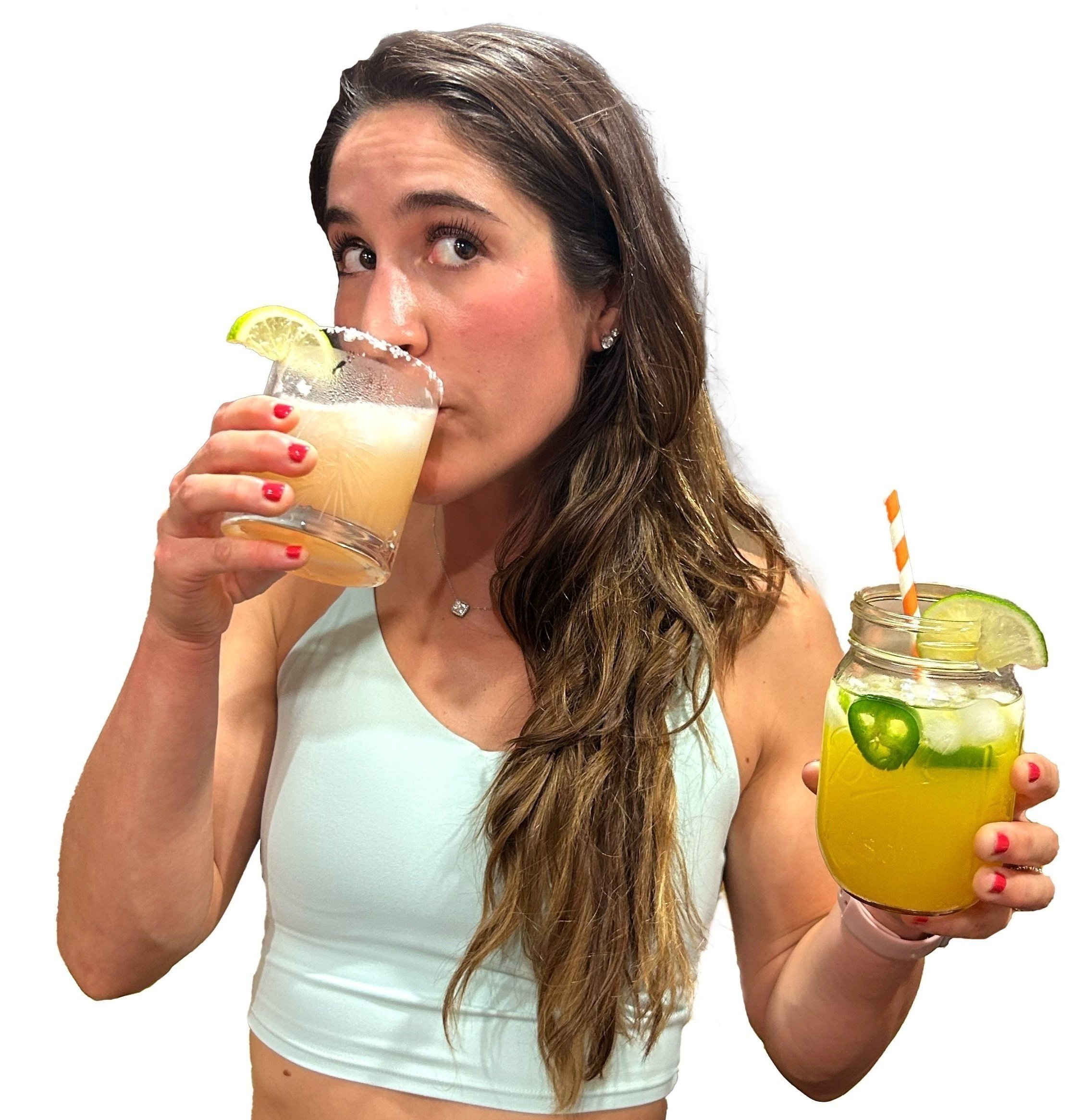 A woman with long wavy brown hair drinking a cocktail with a lime wedge garnished on the glass. She is holding another mason jar-style glass with a yellow-green drink containing lime and jalapeno slices, with a striped straw. She is dressed in a white sleeveless top and has red painted nails.