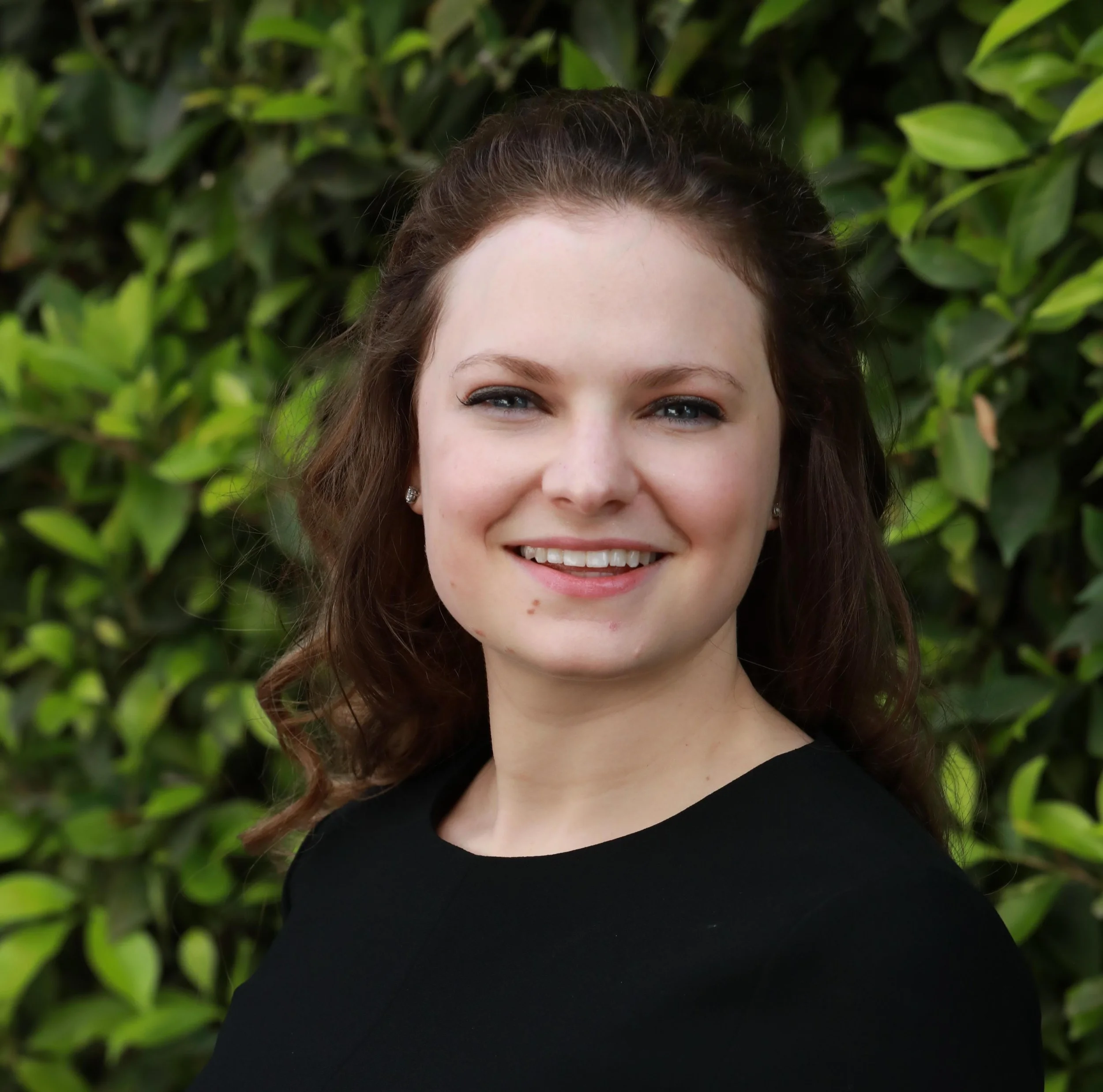 A young woman with brown hair, blue eyes, and earrings, smiling outdoors in front of green foliage.