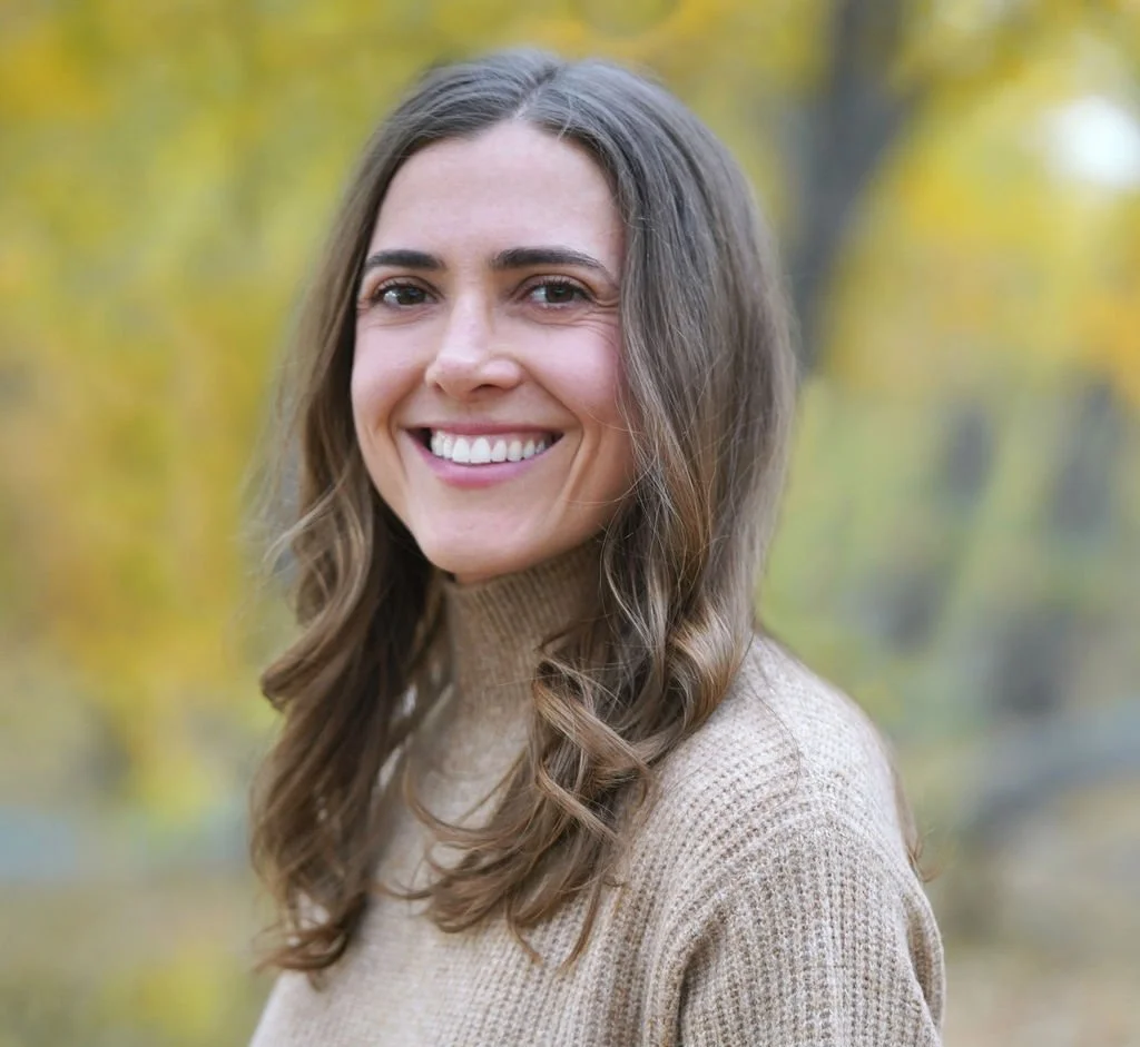 A woman with long, wavy brown hair smiling outdoors in front of trees with yellow and green leaves.