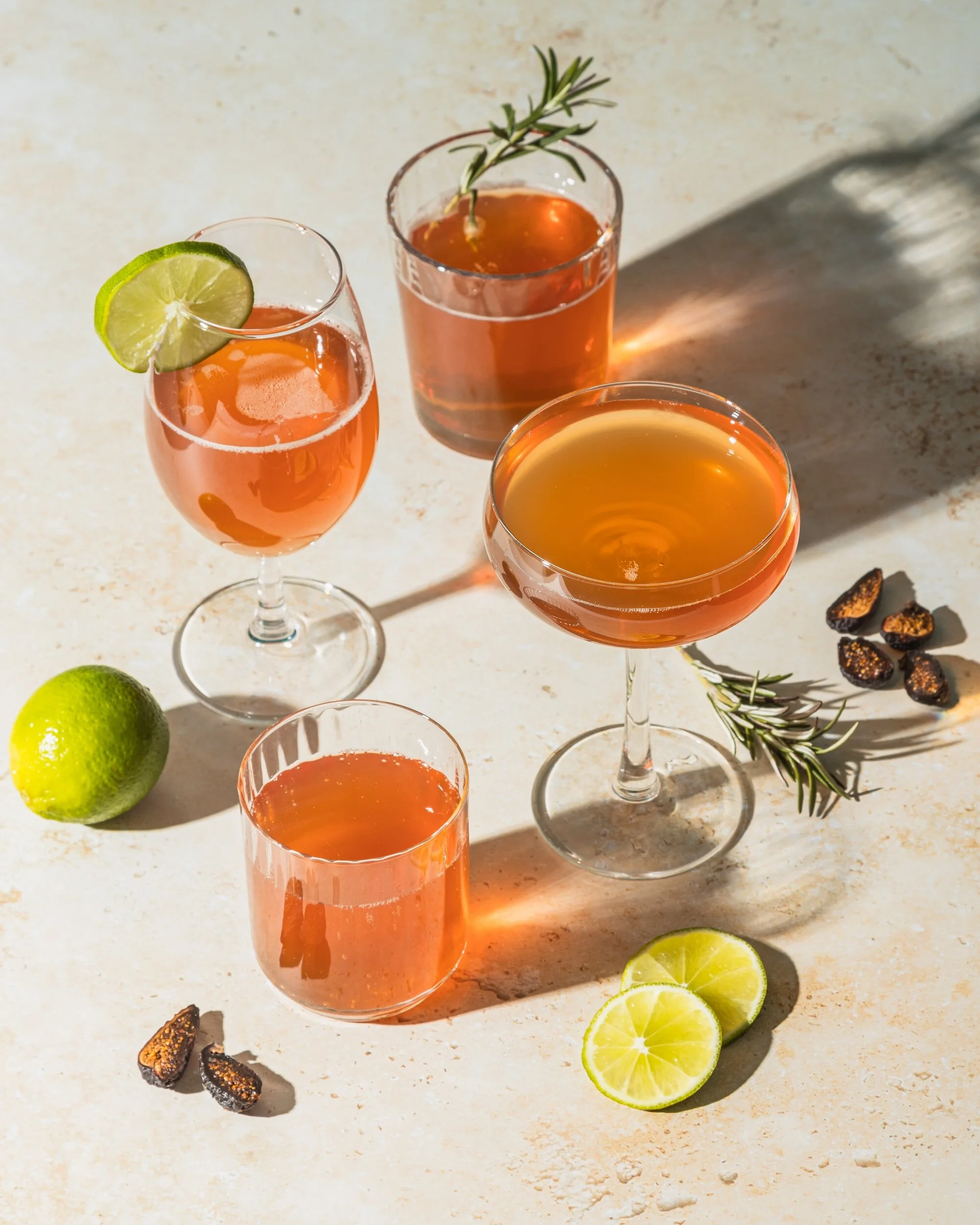 Four glasses of cocktails with lime slices, rosemary sprigs, and dates surrounding them on a light-colored surface.