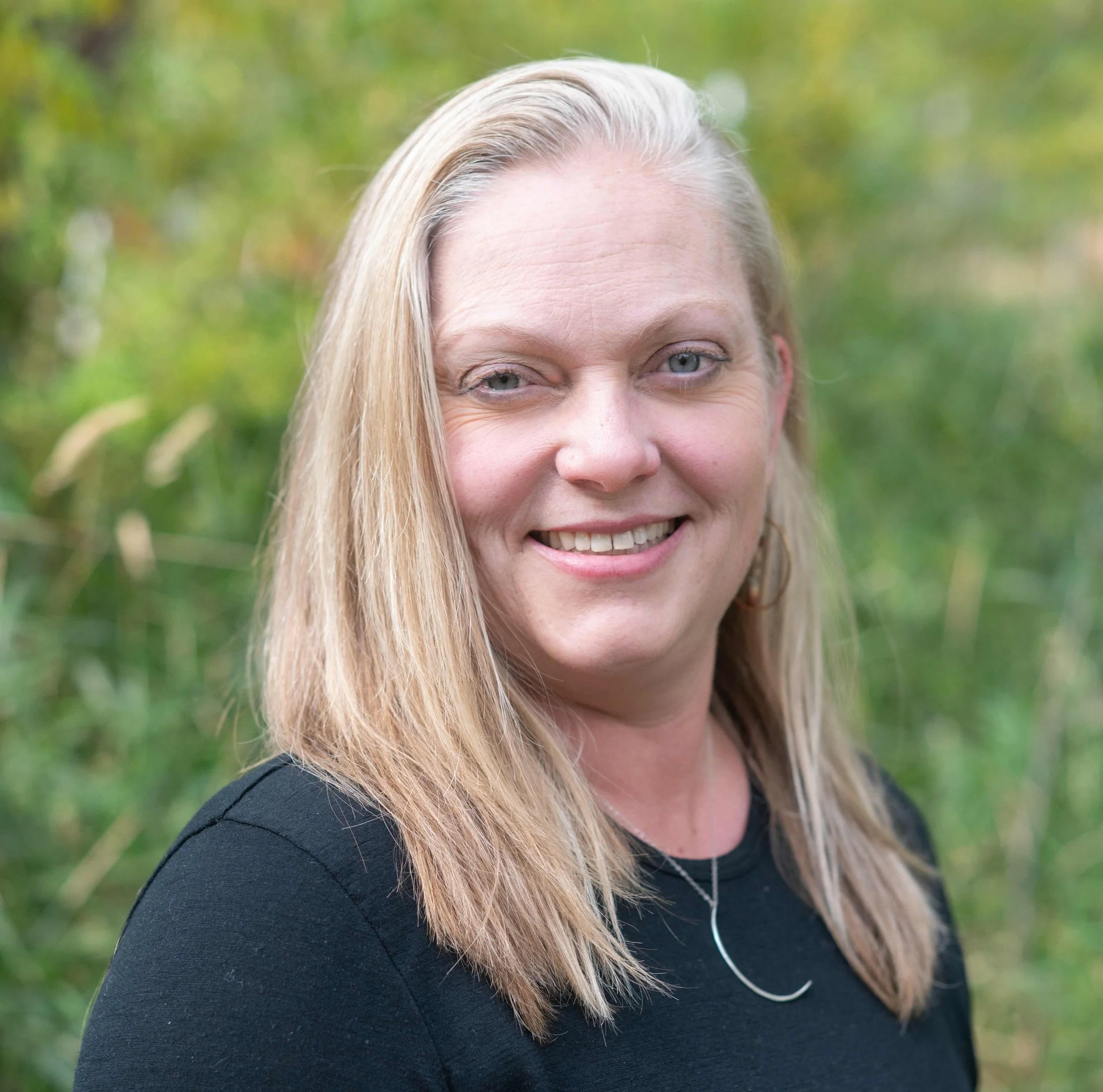 A woman with blonde hair and a black top smiling outdoors with green foliage in the background.