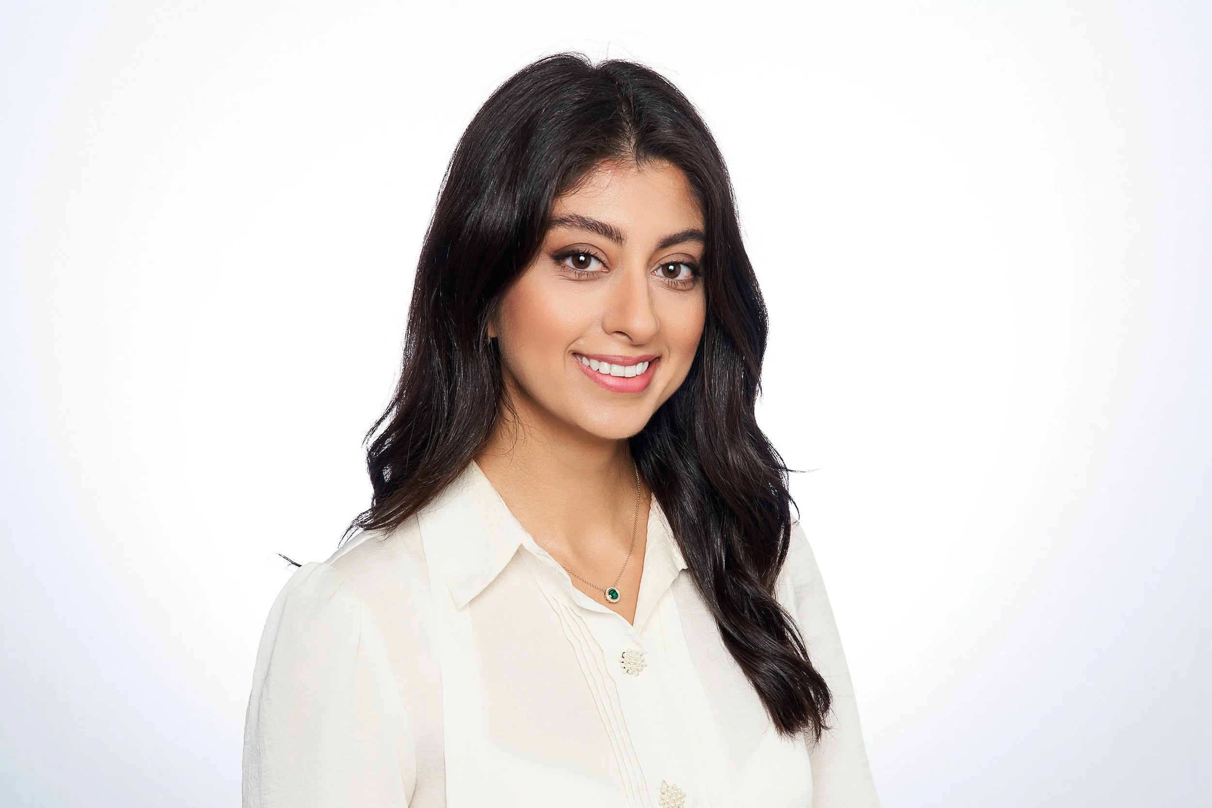 A young woman with long dark hair wearing a white blouse and a necklace with a green gemstone, smiling against a plain white background.