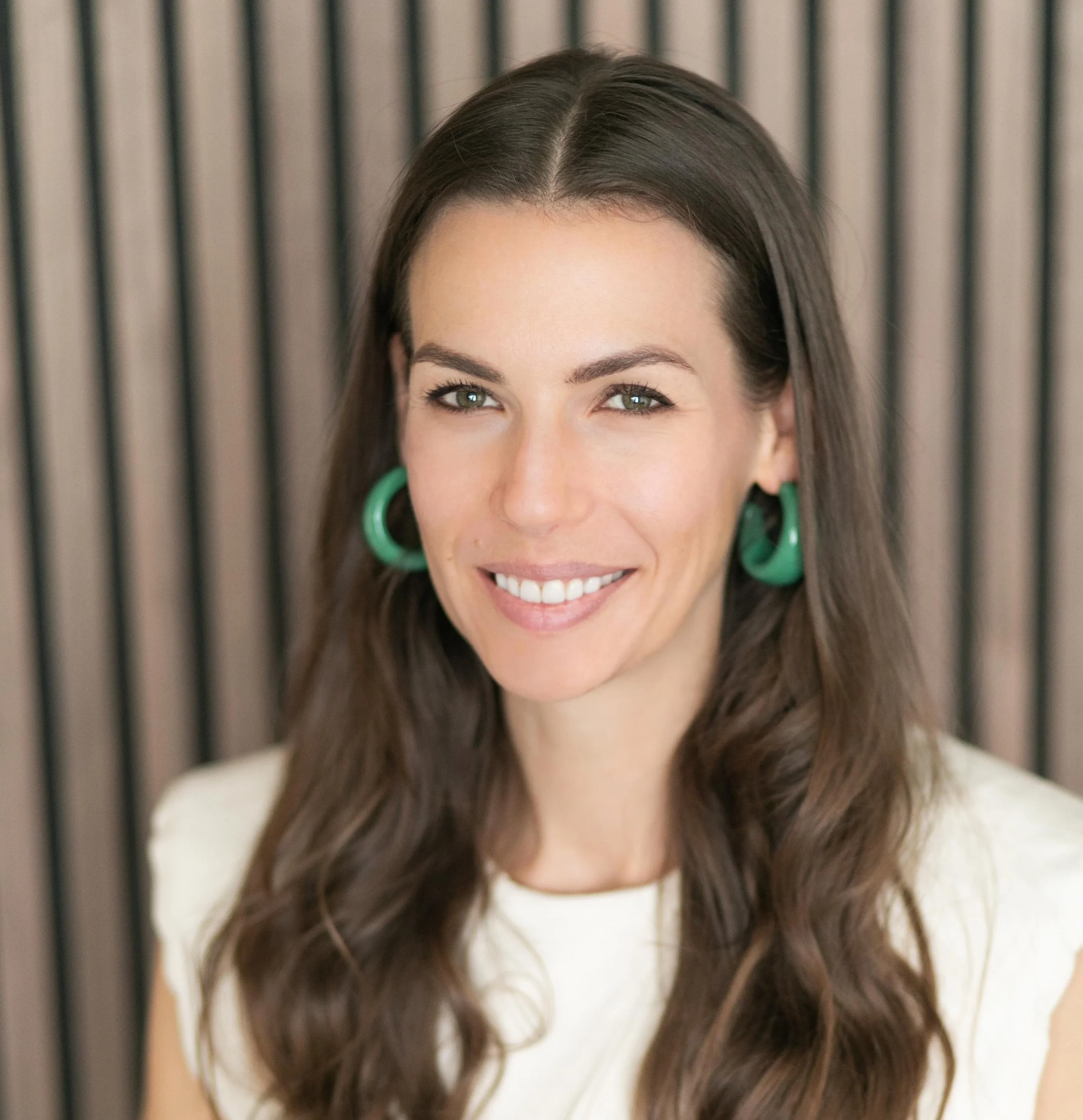 A woman with long brown hair, green earrings, and a light-colored top smiling at the camera, with vertical wooden slats in the background.