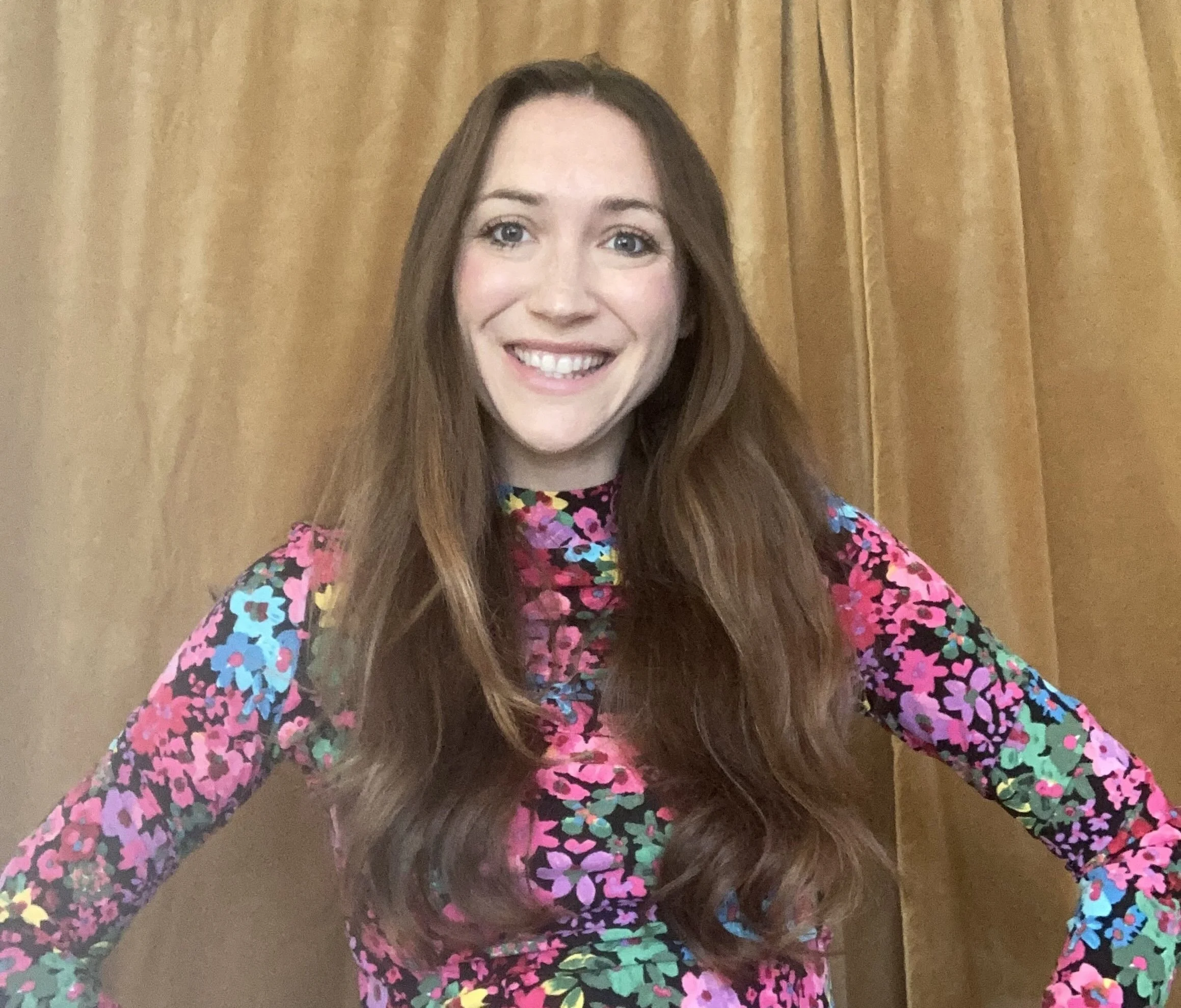 A woman with long brown hair smiling, wearing a colorful floral long-sleeve top, standing in front of a gold curtain.
