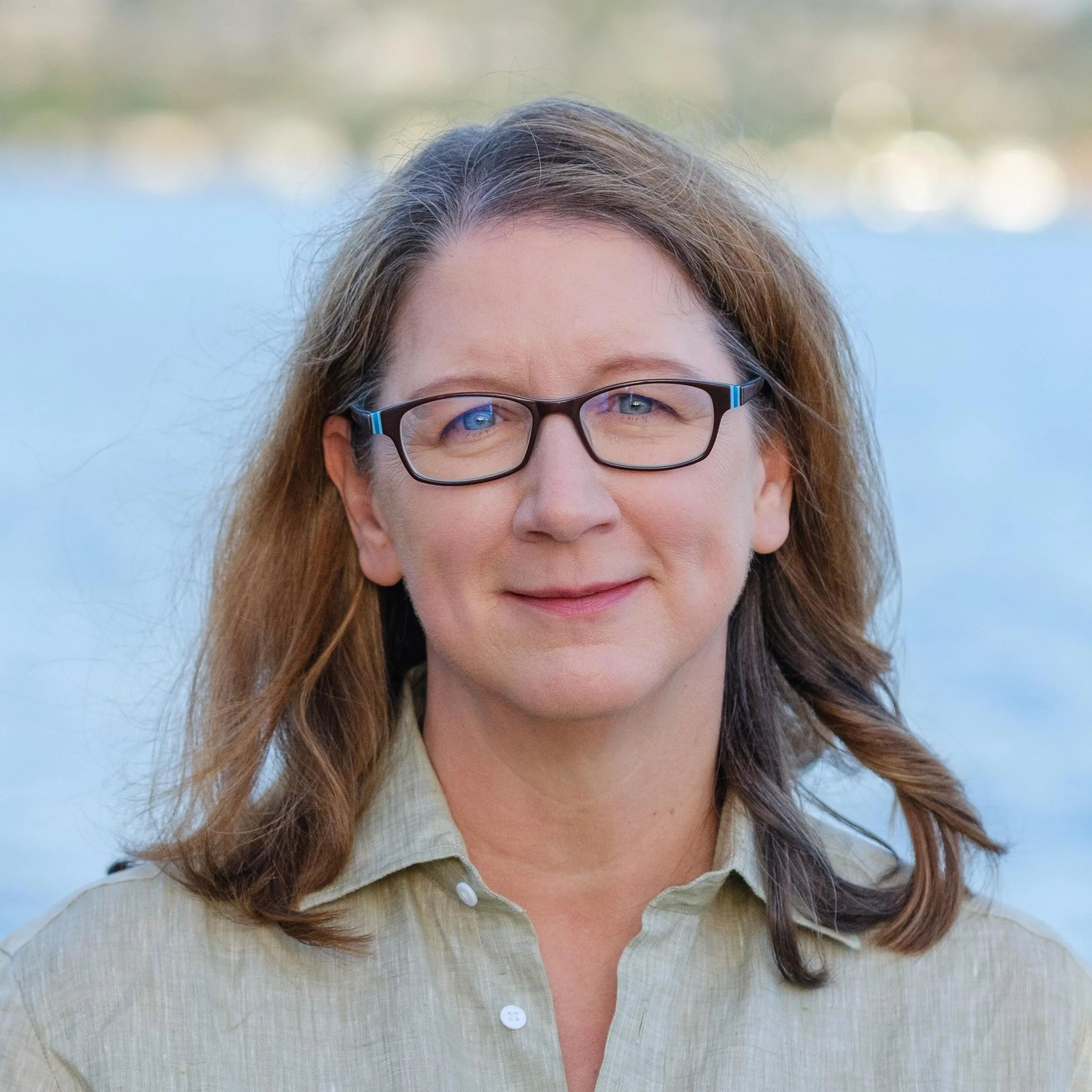 A woman with brown hair, glasses, and blue eyes, wearing a beige button-up shirt, standing outdoors with a body of water in the background.