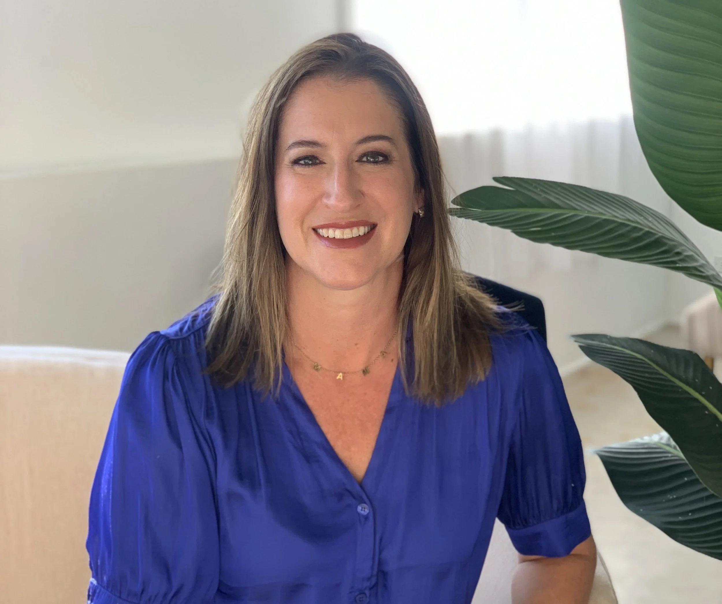A woman with shoulder-length light brown hair wearing a royal blue blouse sitting indoors near a green plant with large leaves, smiling at the camera.