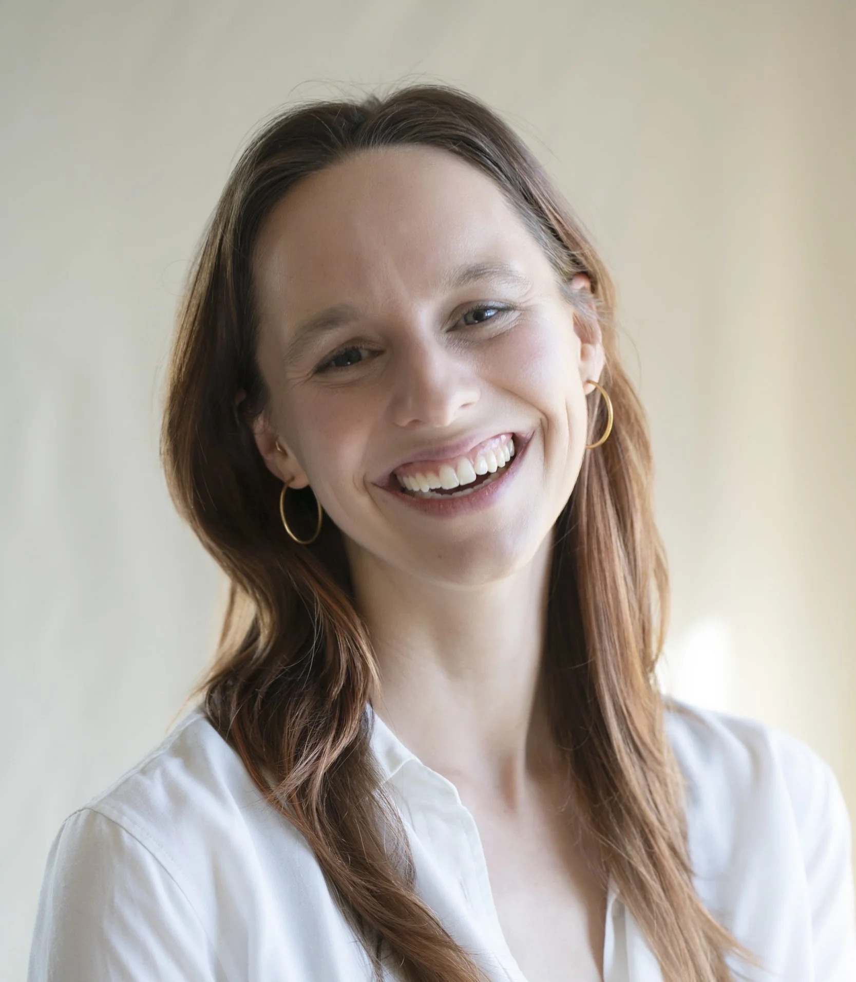 A woman with long, wavy, reddish-brown hair, smiling, wearing a white shirt and gold hoop earrings, standing against a neutral background.
