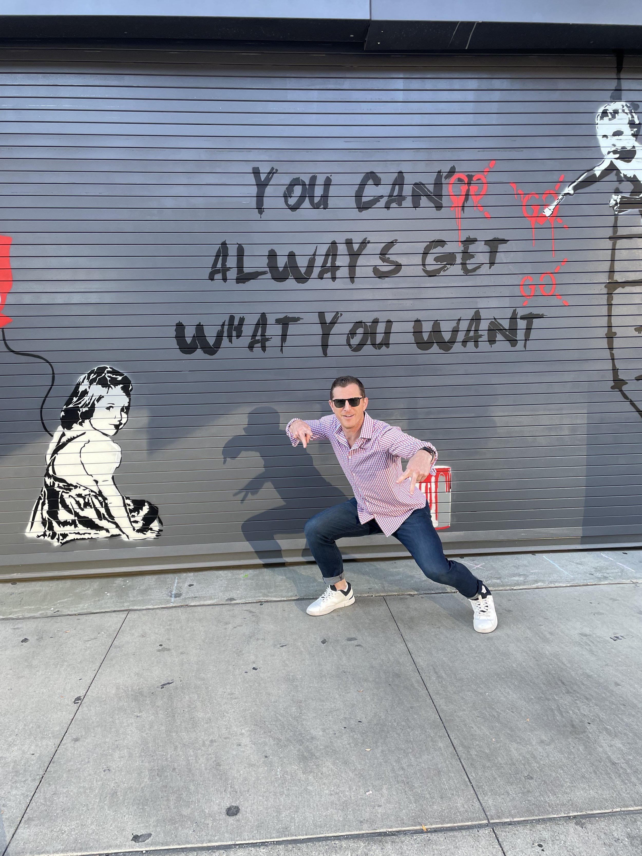 Person posing in front of mural with text "You can't always get what you want" on a gray wall, featuring artwork of two figures and red paint accents.