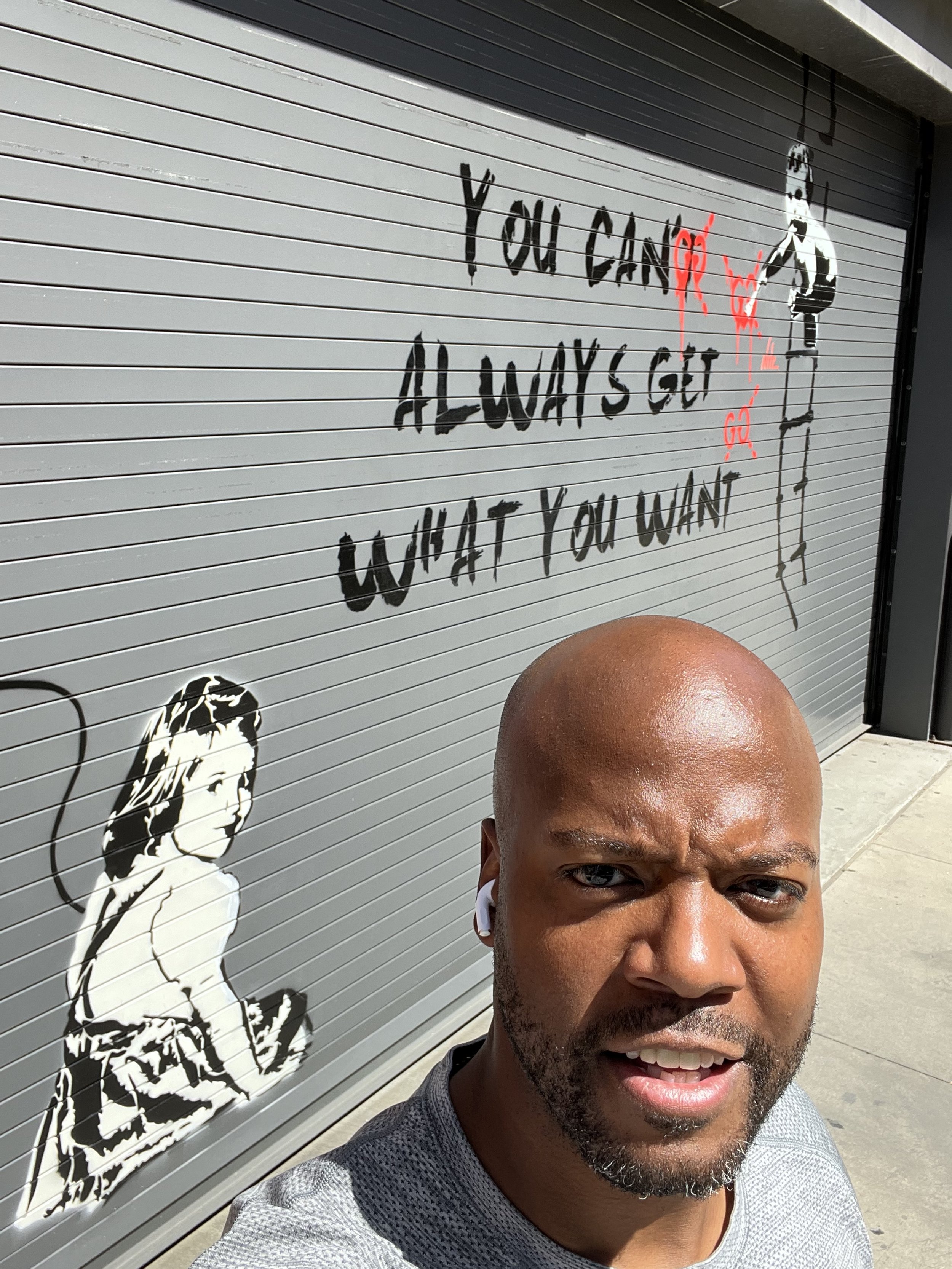 A man taking a selfie in front of a gray wall with graffiti text saying "You Can't Always Get What You Want" and images of two stenciled figures, one resembling a girl with a skipping rope and another a man holding a spray can, with red paint drips o