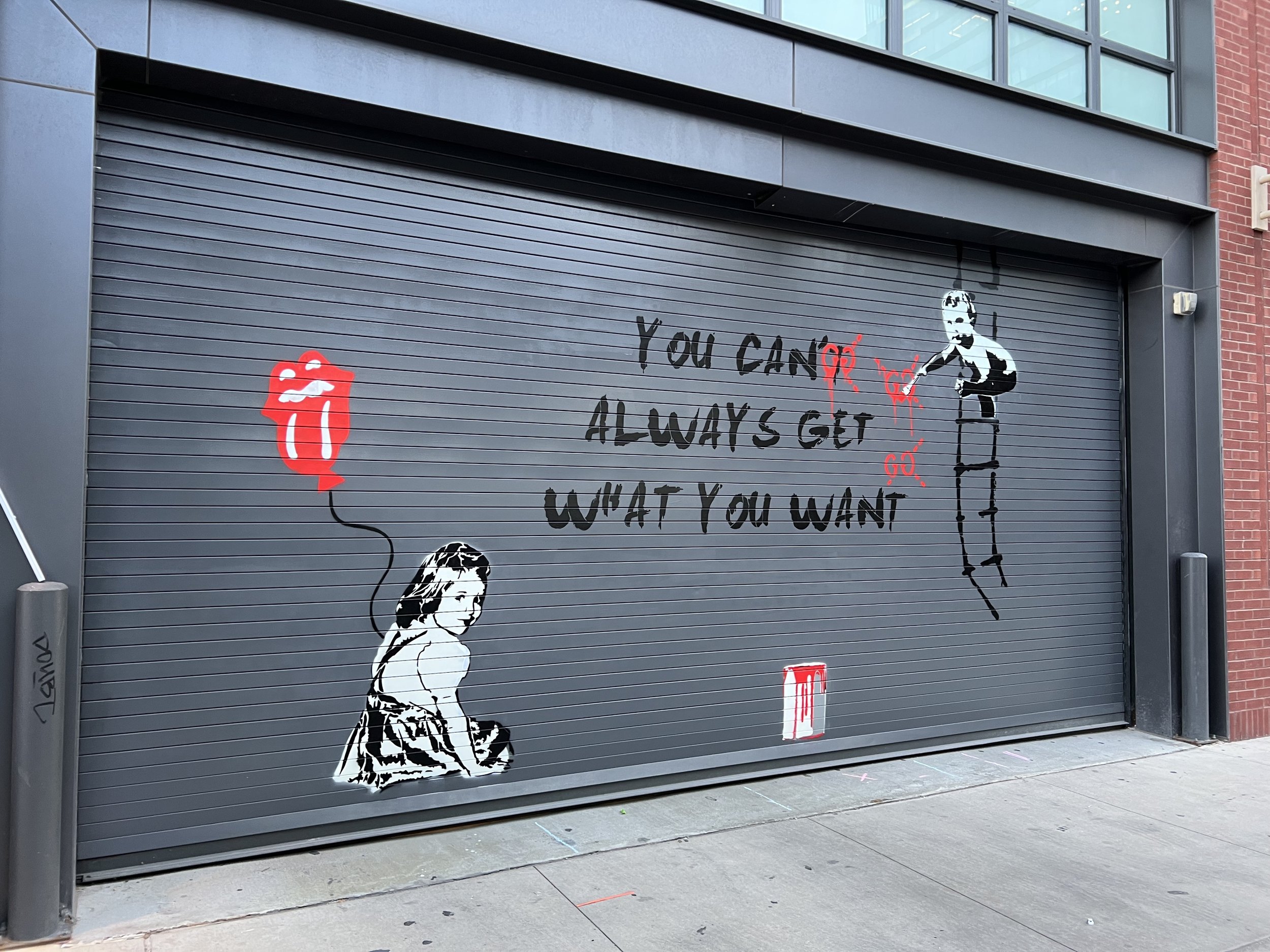 Street art on a garage door with a stenciled girl holding a red balloon shaped like lips and text reading 'You Can't Always Get What You Want.'
