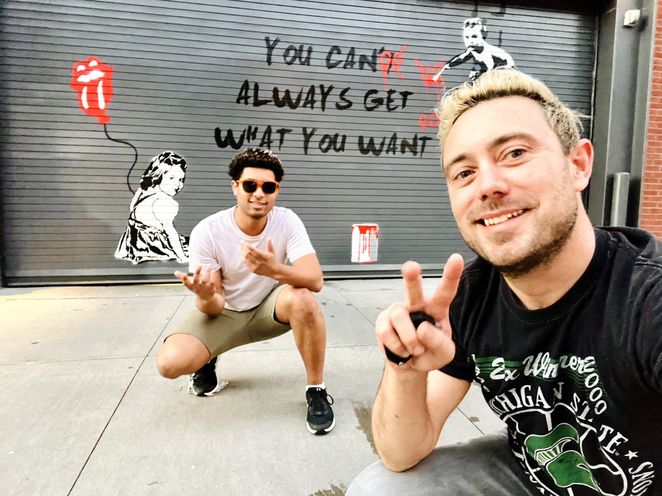 Two people posing in front of a garage door featuring street art. The artwork includes a stencil of a child holding a balloon shaped like a red tongue and lips, and a phrase "You can't always get what you want" painted on the door. One person kneels