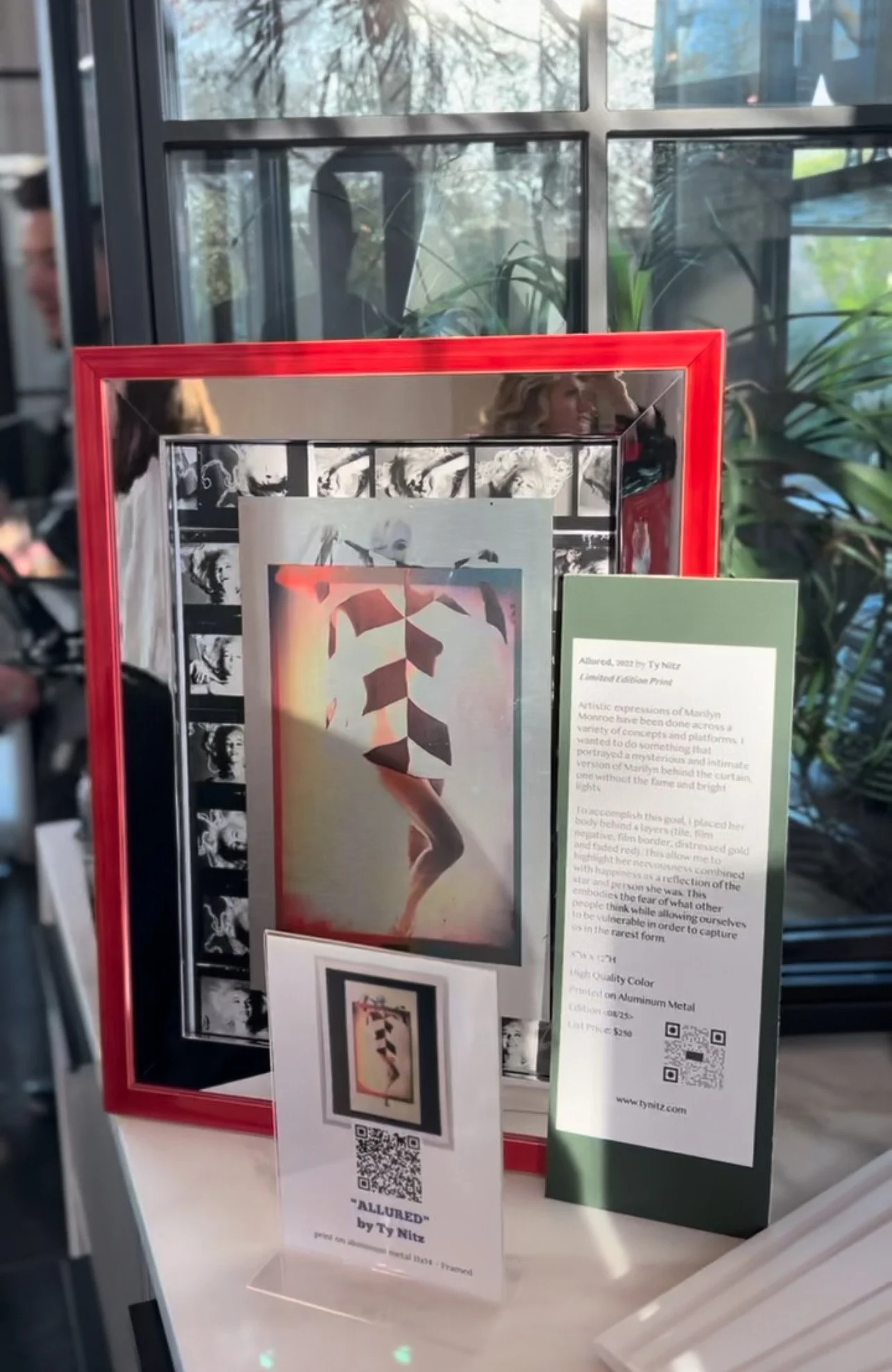 Framed artwork by Ty Nitz titled 'Allured', featuring geometric shapes and a stylized figure. Displayed with informational text and a QR code on a table.