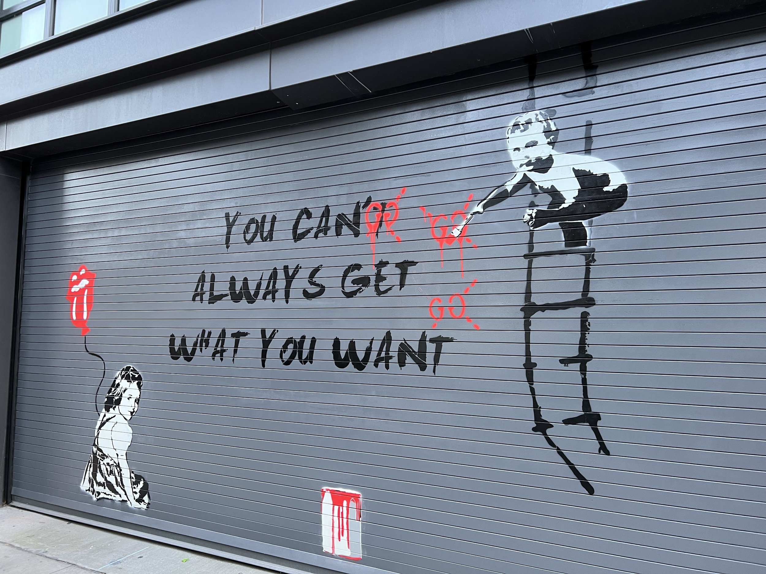 Street art on a gray garage door featuring black text "You can't always get what you want" with red paint splatter and a painted Rolling Stones tongue logo balloon held by a woman. A stencil of a child on a ladder is also included.