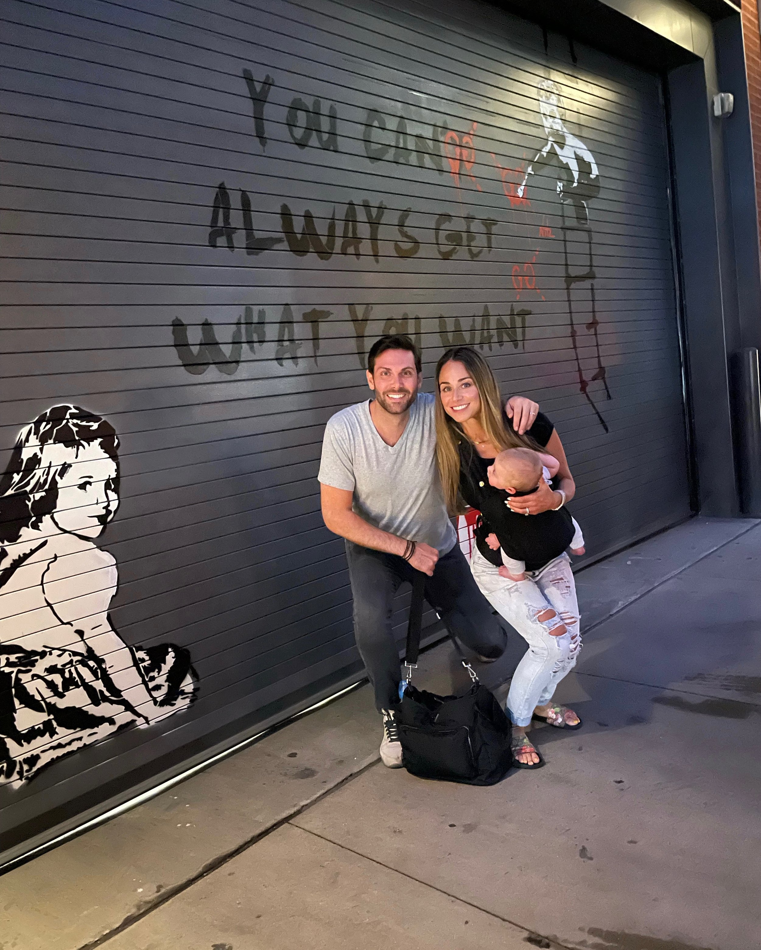 A couple and a baby pose in front of a mural featuring graffiti-style art and text that reads "You can't always get what you want." The mural includes images of a girl and a man. The couple smiles at the camera with their baby.