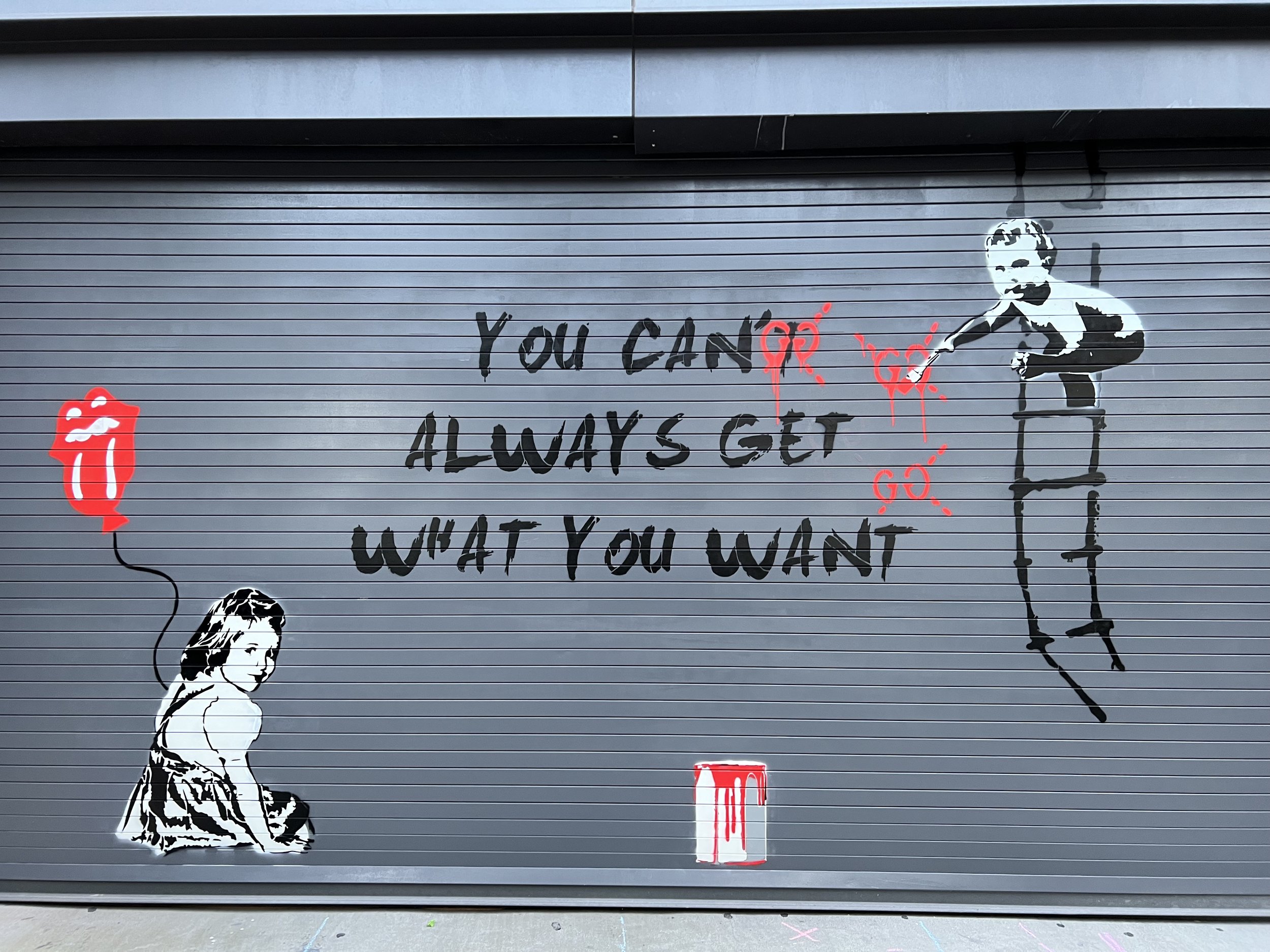 Street art on a metal door featuring the phrase "You can't always get what you want." A girl is painted holding a balloon resembling the iconic Rolling Stones tongue logo on the left, and a stencil of a man painting on the wall with splatters of red
