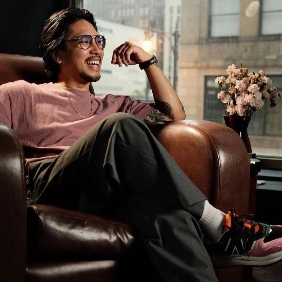 Man sitting comfortably in a leather chair, smiling, wearing glasses, a pink t-shirt, gray pants, and colorful sneakers, with a tattoo on his arm. A vase of pink flowers is on a side table next to him.