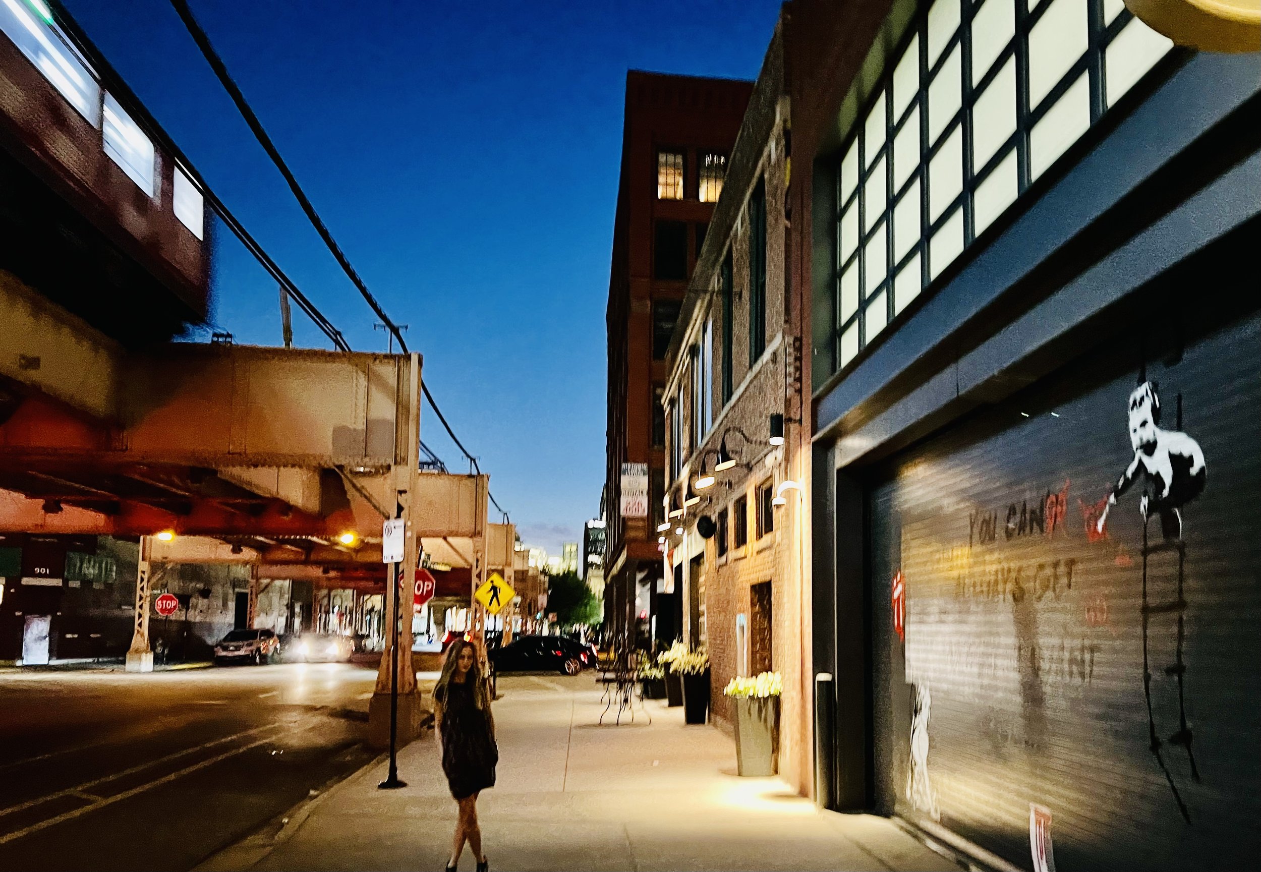 Urban street scene at dusk with elevated train, graffiti art on building, and a person walking on the sidewalk.