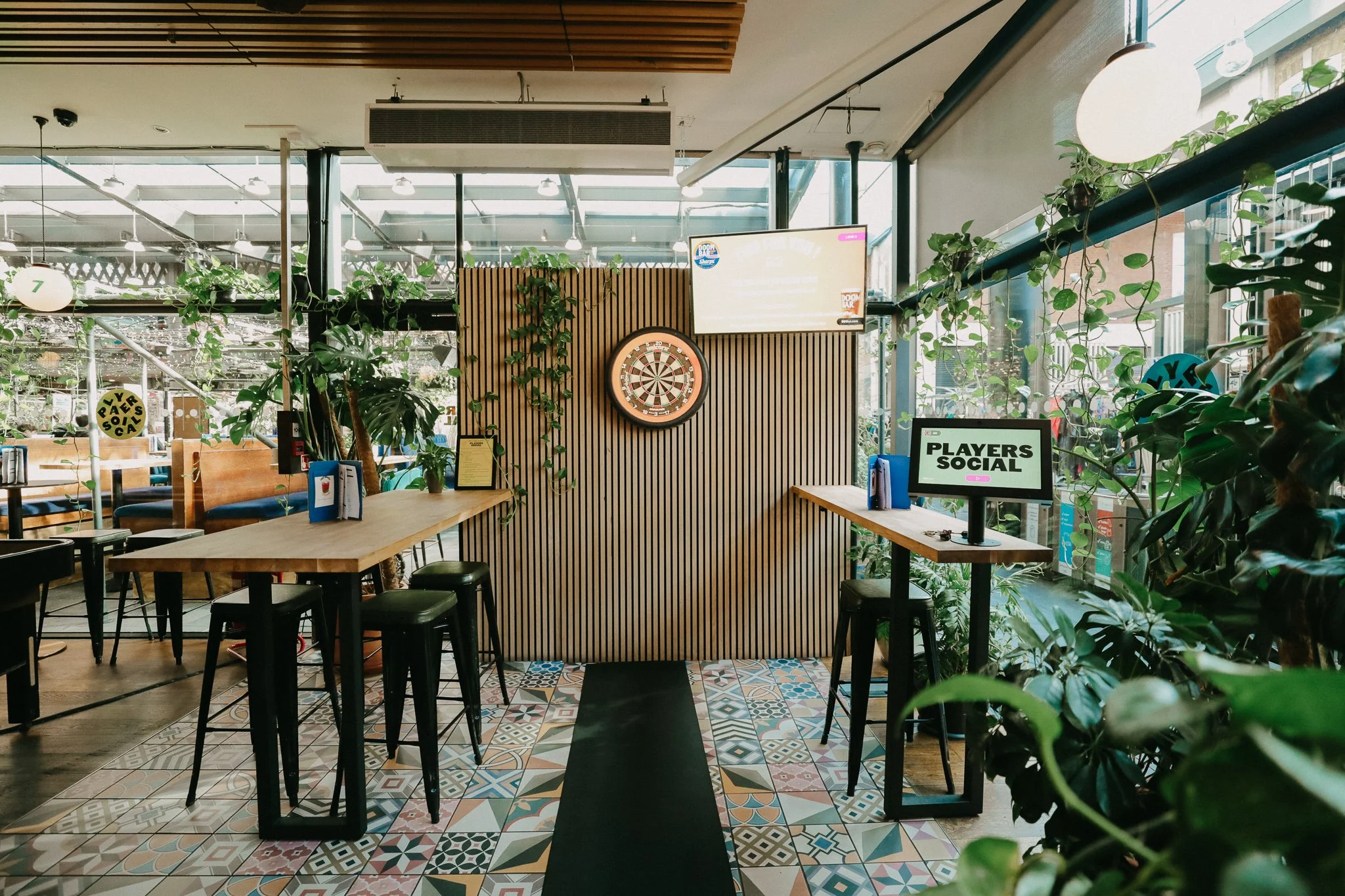 Interior of a modern restaurant or cafe with a central darts game area, decorated with plants, wooden furniture, and patterned tiles on the floor. There are digital screens displaying 'PLAYERS SOCIAL' and a dartboard on a wooden slat wall. 