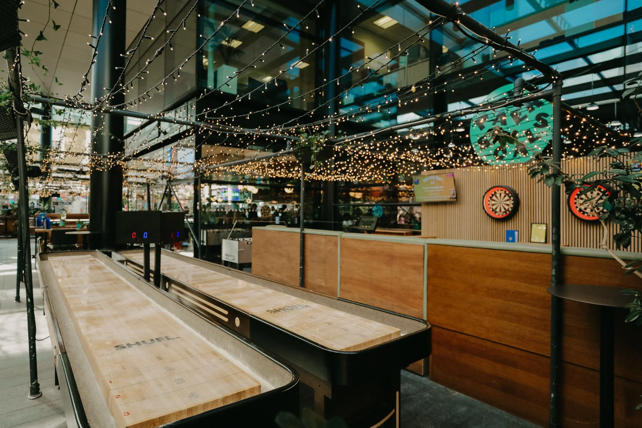 Outdoor shuffleboard court decorated with string lights, with dartboards on the wall in background, in a modern entertainment venue. Players Social Cocktail and Games Bar Spitalfields Market. Cocktail classes in London