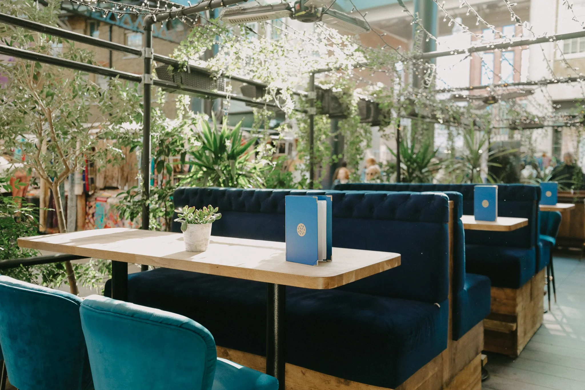 Indoor and outdoor restaurant with blue velvet booth seating, wooden tables, potted plants, and string lights hanging overhead, with a glass ceiling and plants around. Players Social Cocktail and Games Bar Spitalfields Market London