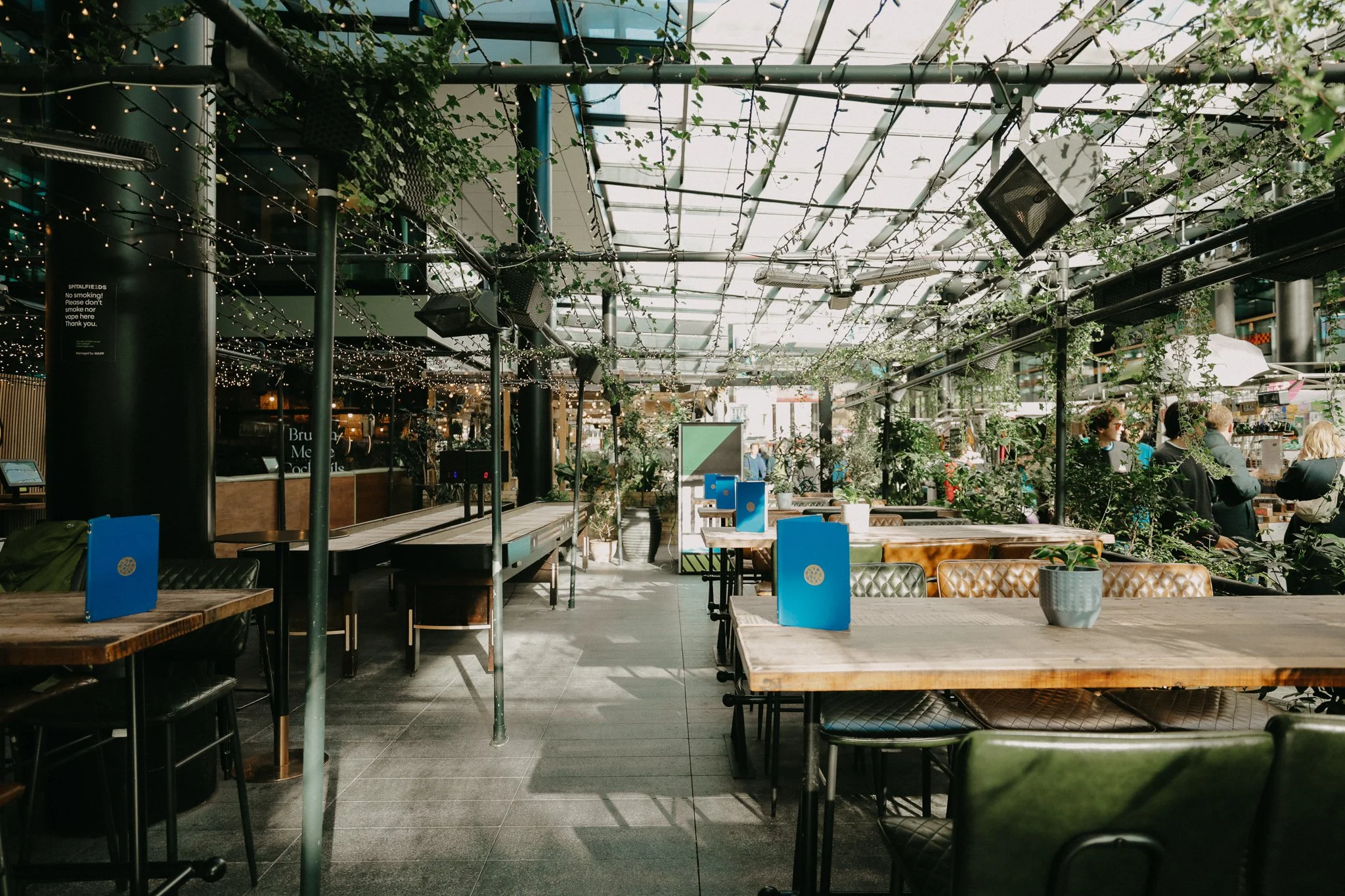 Outdoor view of a modern venue with wooden tables, greenery, and string lights, outdoor seating area with a glass roof. Players Social Cocktail and Games Bar Spitalfields Market. Cocktail classes in London
