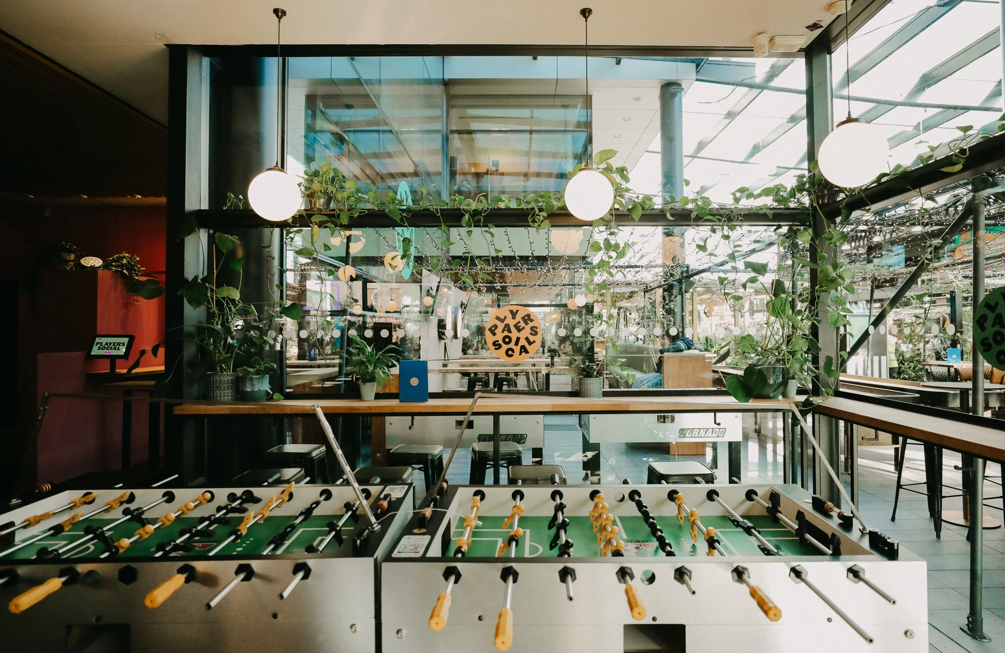 Indoor area of a modern cafe or lounge with large windows, plants, and string lights, featuring a foosball table in the foreground. Players Social Cocktail and Games Bar Spitalfields Market. Cocktail classes in London