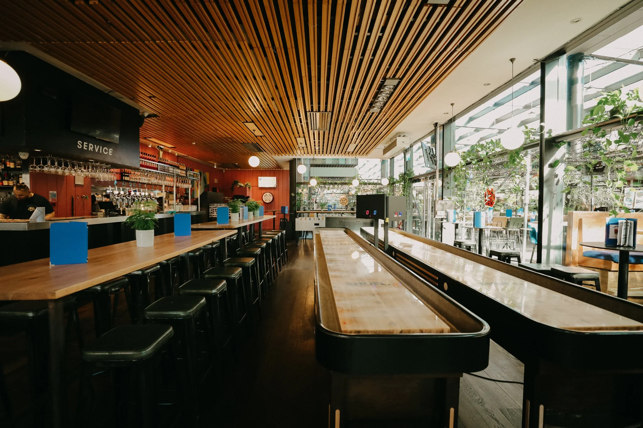 Empty bar and restaurant interior with a long wooden counter, bar stools, and large windows with plants outside, decorated with hanging globe lights. Players Social Cocktail and Games Bar Spitalfields Market. Cocktail classes in London