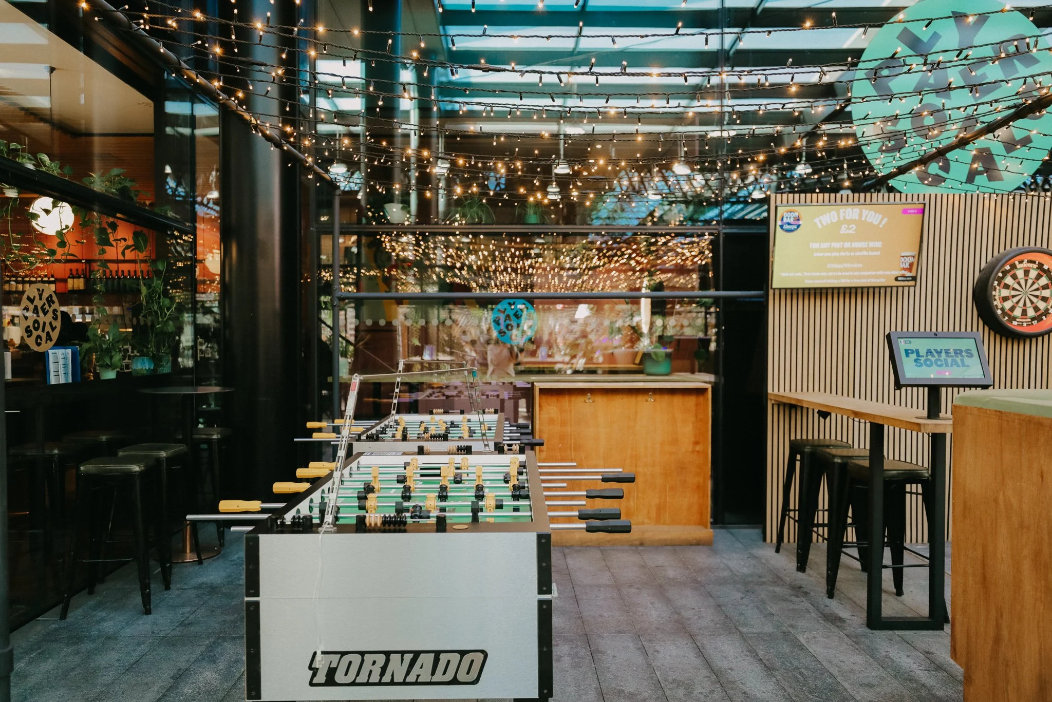 Outdoor dartboard area with a foosball table, a bar counter, string lights above, and signs that say 'Players Social'. Players Social Cocktail and Games Bar Spitalfields Market. Cocktail classes in London
