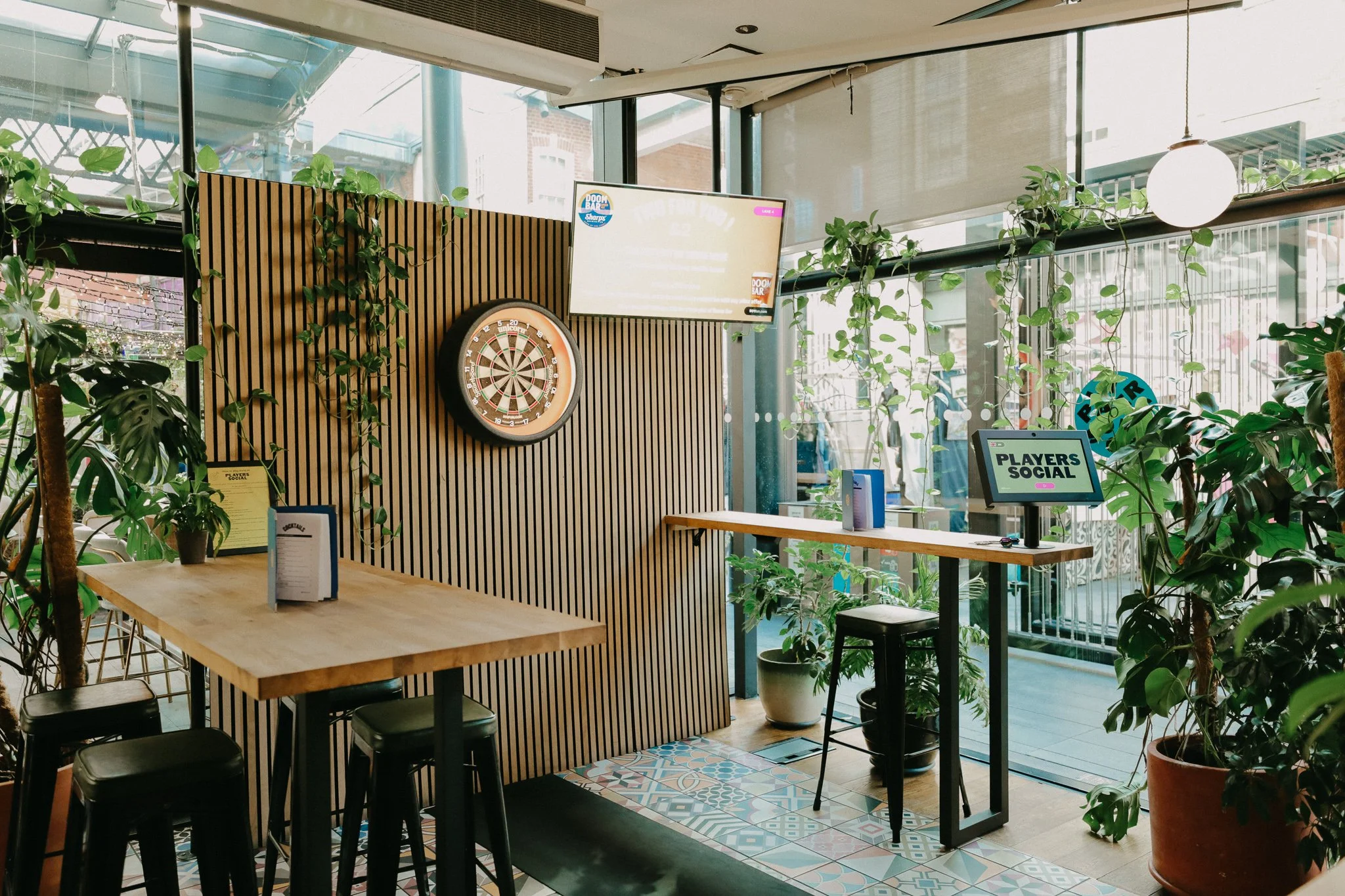Interior of a modern bar with wooden tables, potted plants, a dartboard on a wooden slat wall, and digital screens displaying social and event information. Large windows let in natural light. Players Social Cocktail and Games Bar Spitalfields Market.