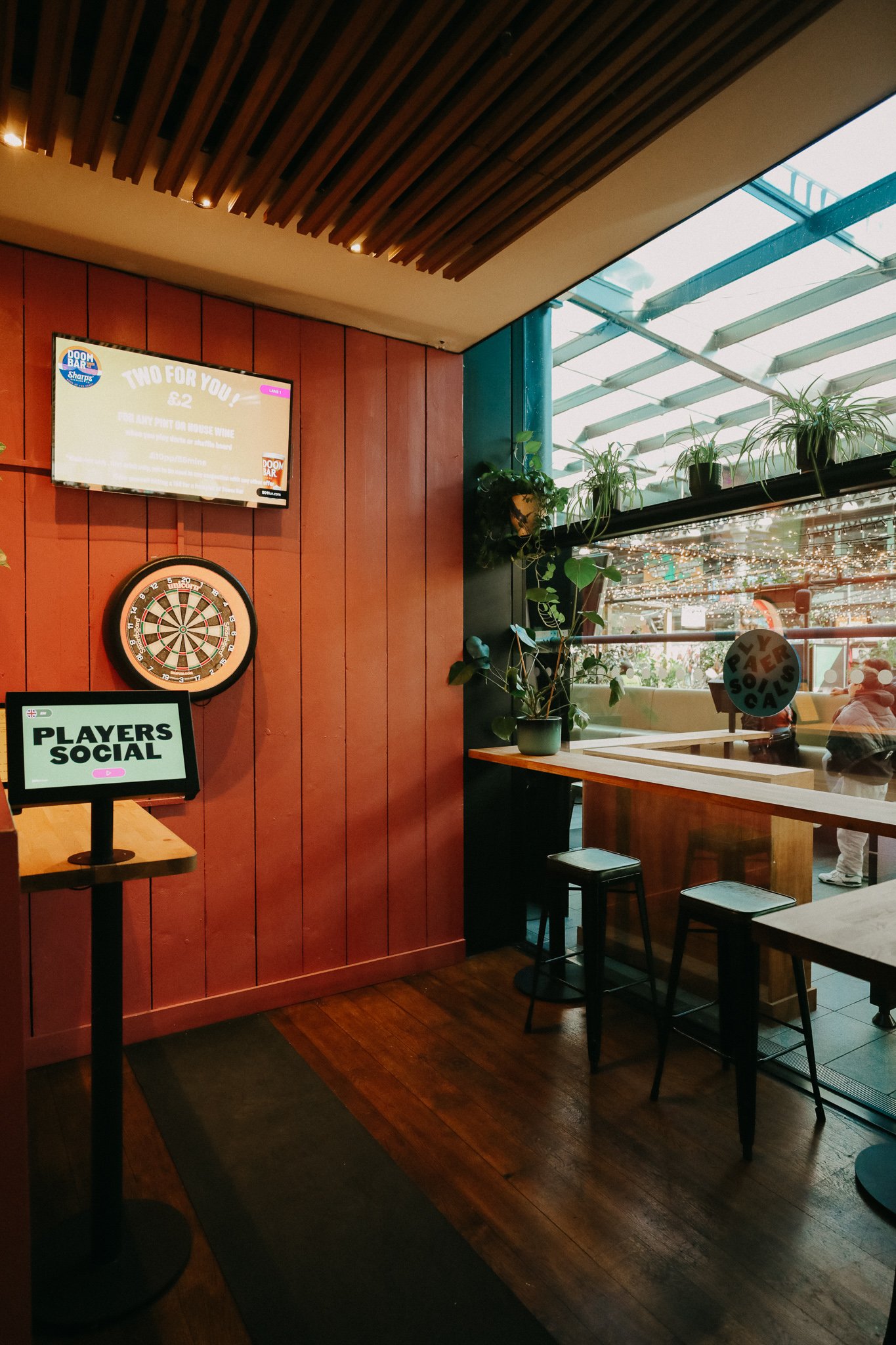 Indoor space with wooden flooring and walls, decorated with plants, a dartboard, a television, and a sign that reads 'Players Social'. Large glass windows show a seating area outside with people and string lights. Players Social Cocktail & Games Bar