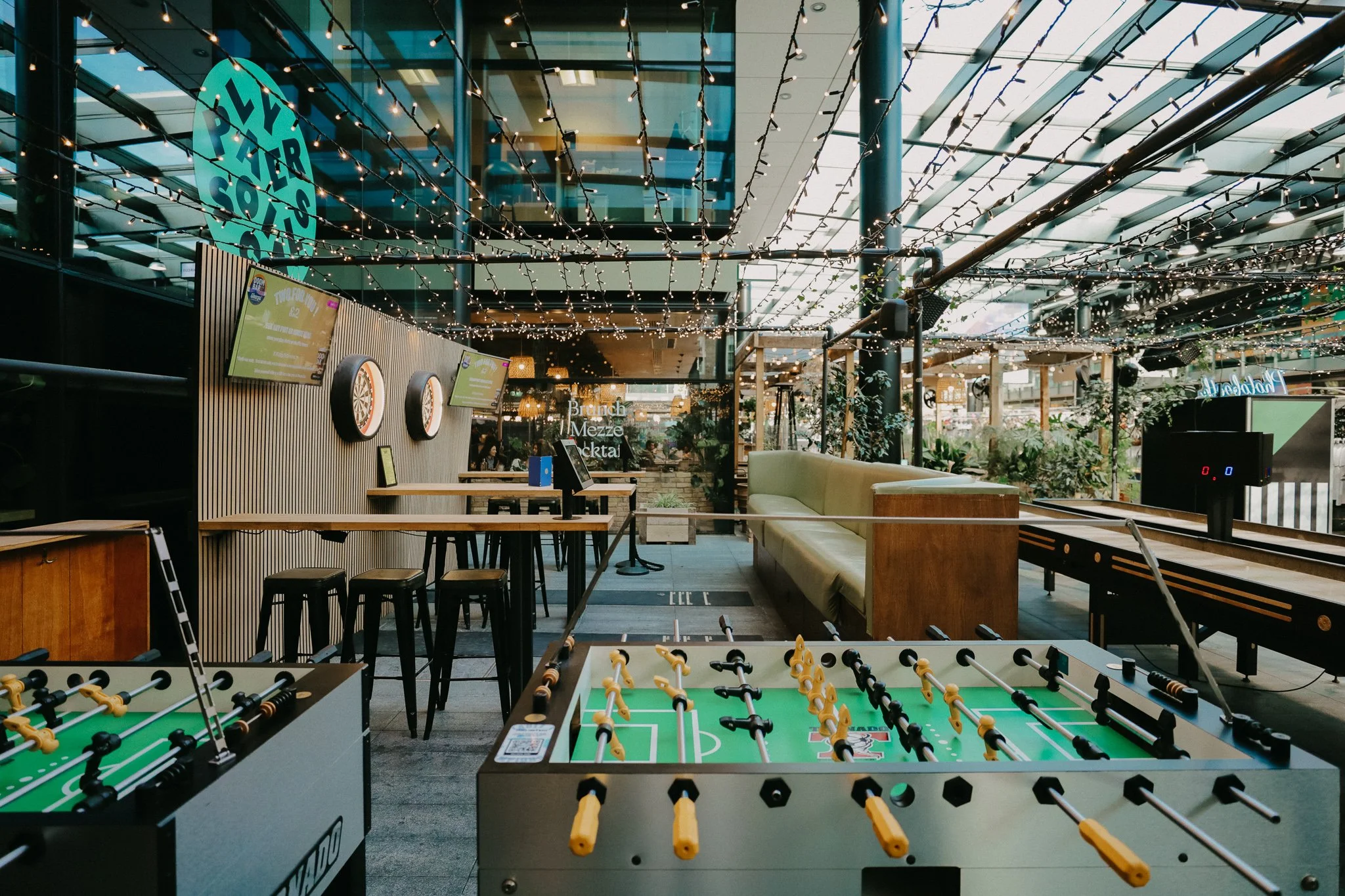 Outdoor recreational area with foosball tables, high tables with stools, a cushioned bench, string lights overhead, a dartboard on the wall, and a bar or seating area in the background with plants and windows. Players Social London