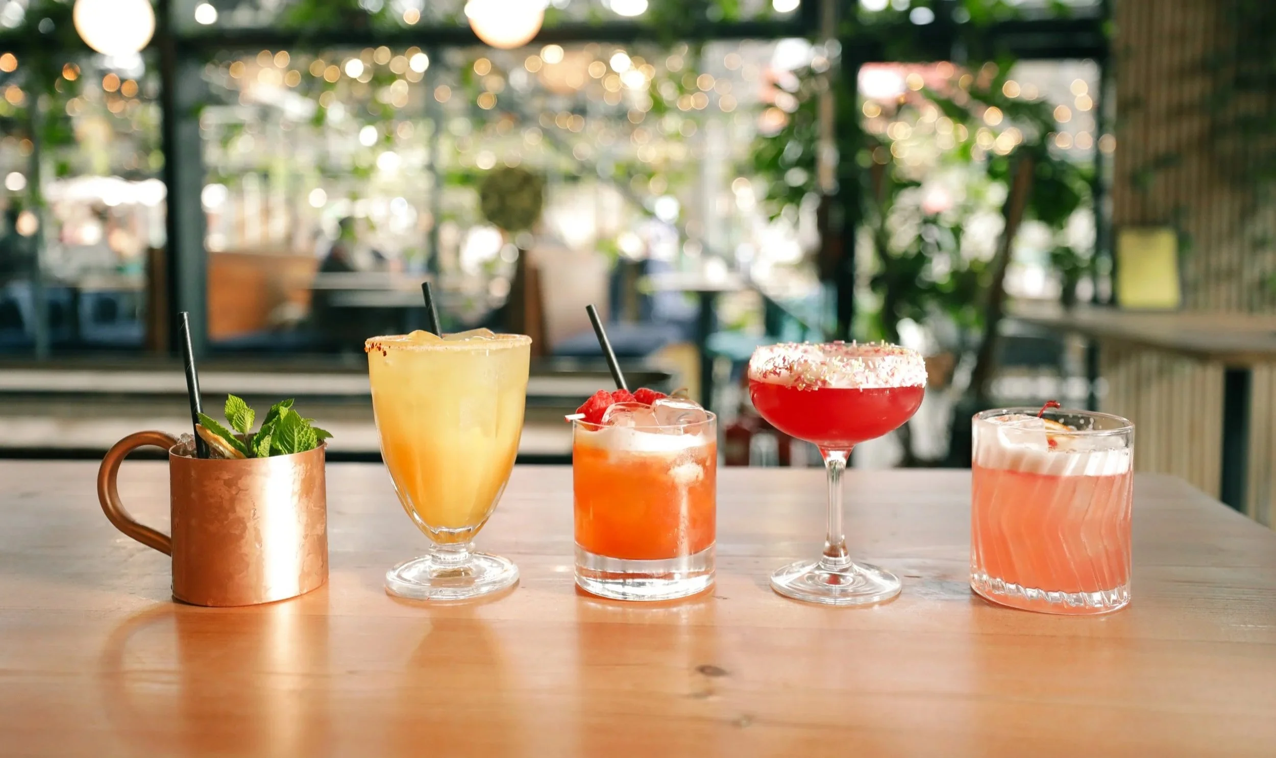 Five colorful cocktails on a wooden table in a bright indoor setting. Players Social Cocktail and Games Bar Spitalfields Market. Cocktail classes in London