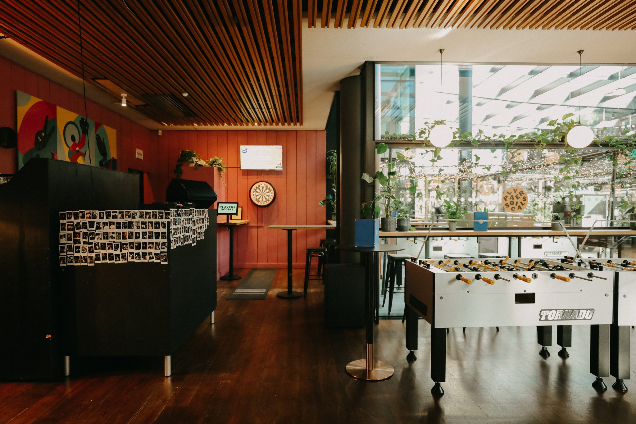 Indoor recreational space featuring a foosball table, a dartboard, and a wall decorated with small photos, with natural light streaming in through large windows and ceiling lights, plants, and artwork on the walls. Players Social Cocktail and Games 