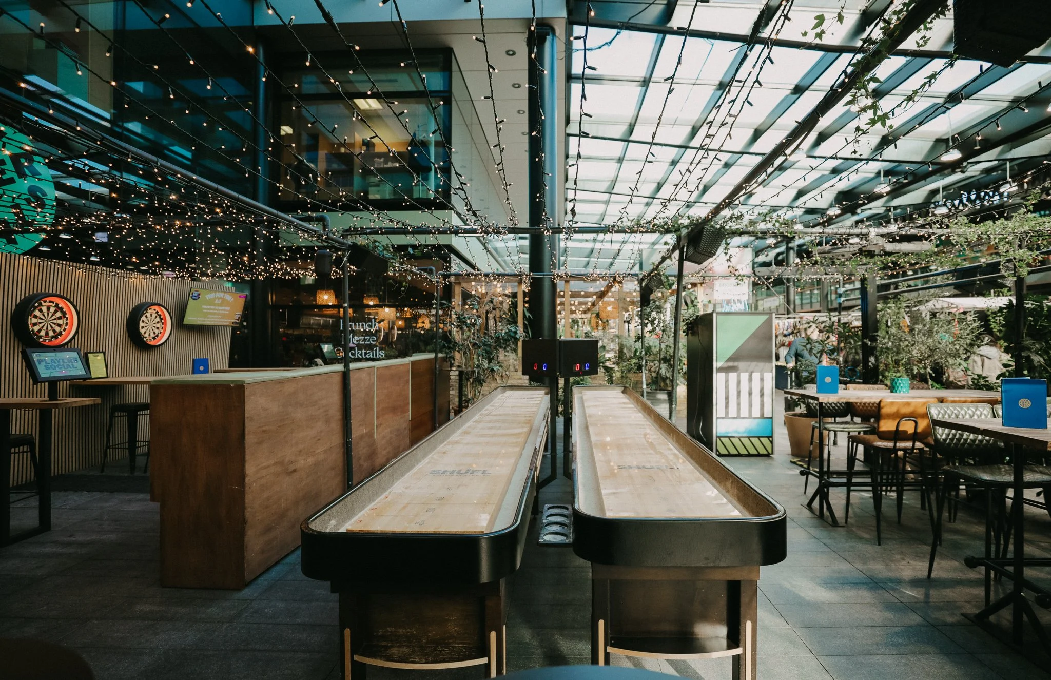 Outdoor bar or restaurant with string lights, dart boards, tables, and ice skating rink in the centre. Players Social Cocktail and Games Bar Spitalfields Market. Cocktail classes in London
