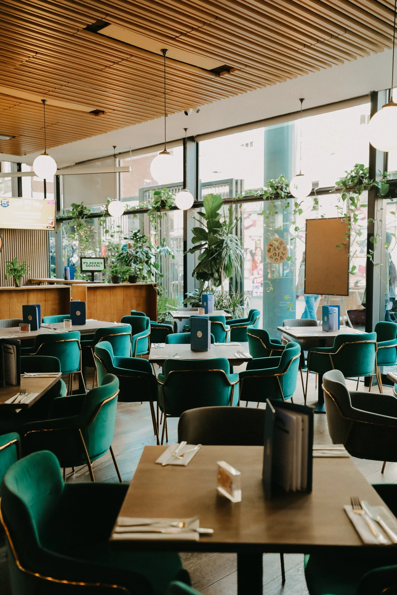 Interior of a modern restaurant with teal chairs, wooden tables set with silverware, and large windows with plants outside, decorated with round hanging lights. Players Social Cocktail and Games Bar Spitalfields Market. Cocktail classes in London