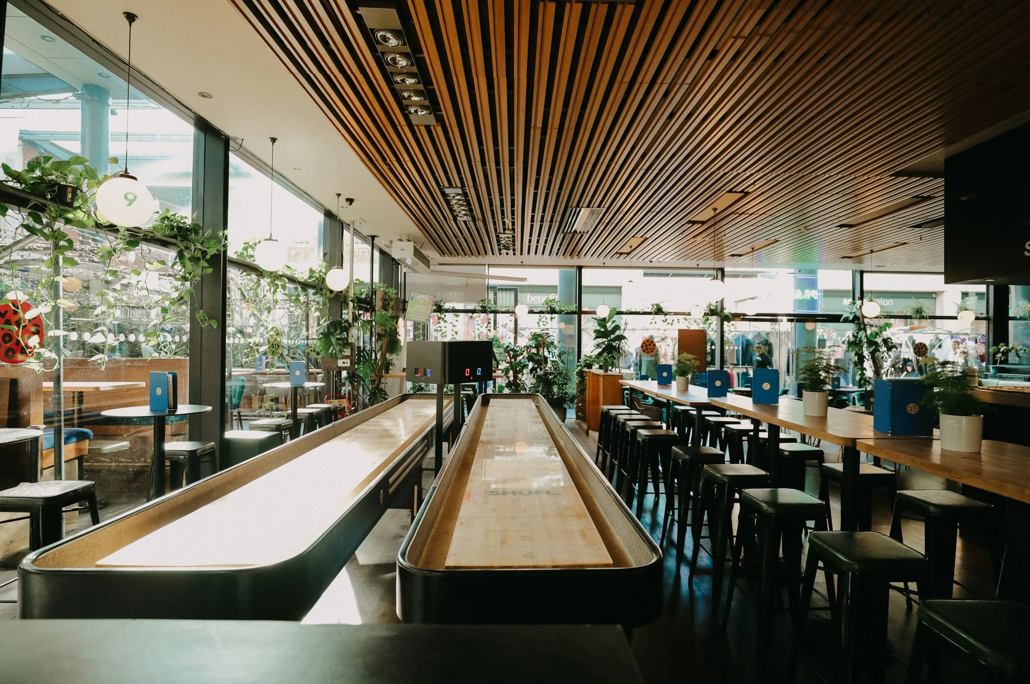 Empty indoor recreational space with shuffleboard courts, bar stools, and large windows with green plants, in a modern, well-lit setting. Players Social Cocktail and Games Bar Spitalfields Market. Cocktail classes in London