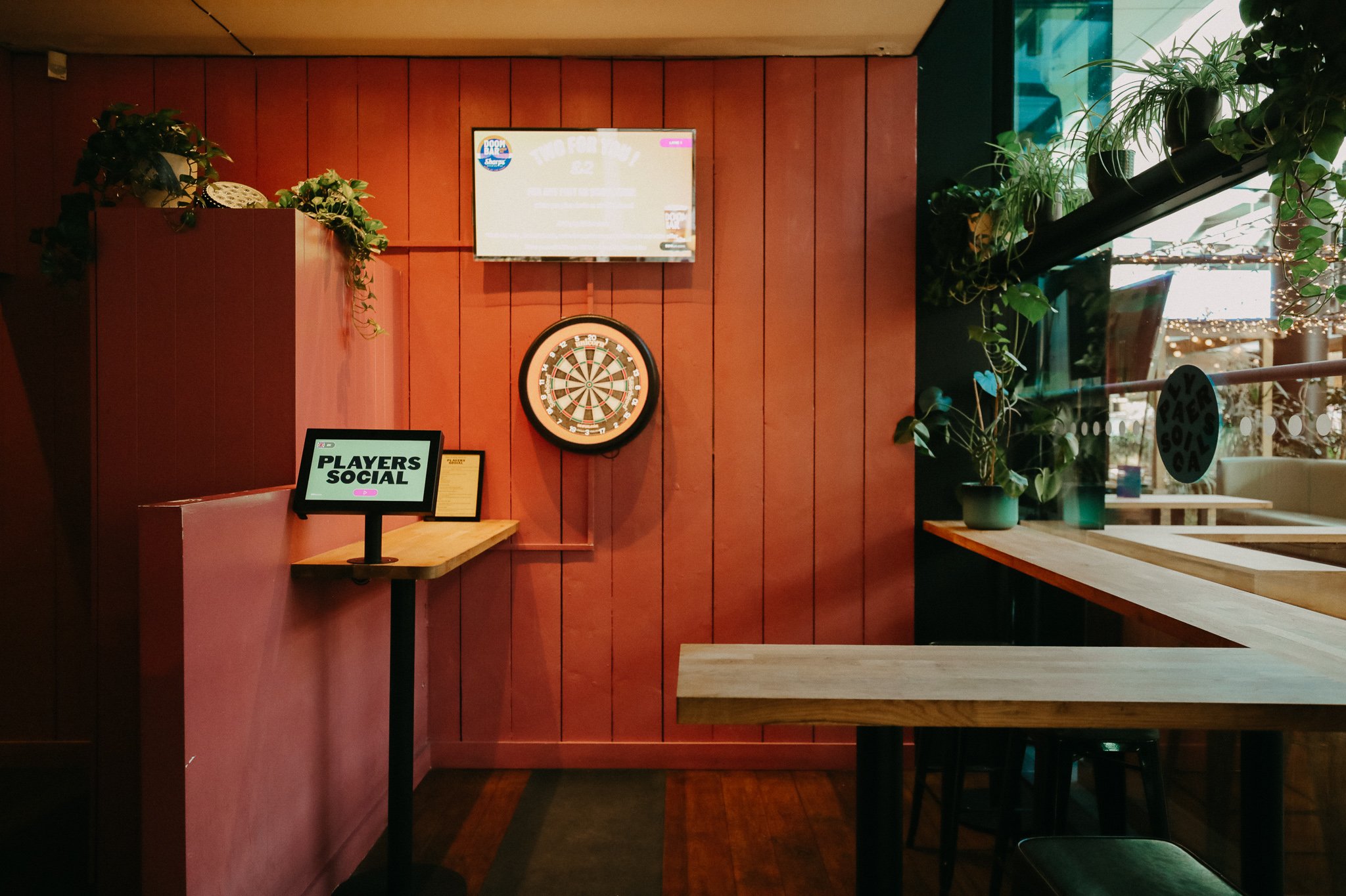 Interior of a pub or bar with a dartboard on a pink wooden wall, a small digital sign on a stand reading 'PLAYERS SOCIAL,' potted plants on a wooden counter, and a large window showing outdoor lights. Players Social Cocktail and Games Bar