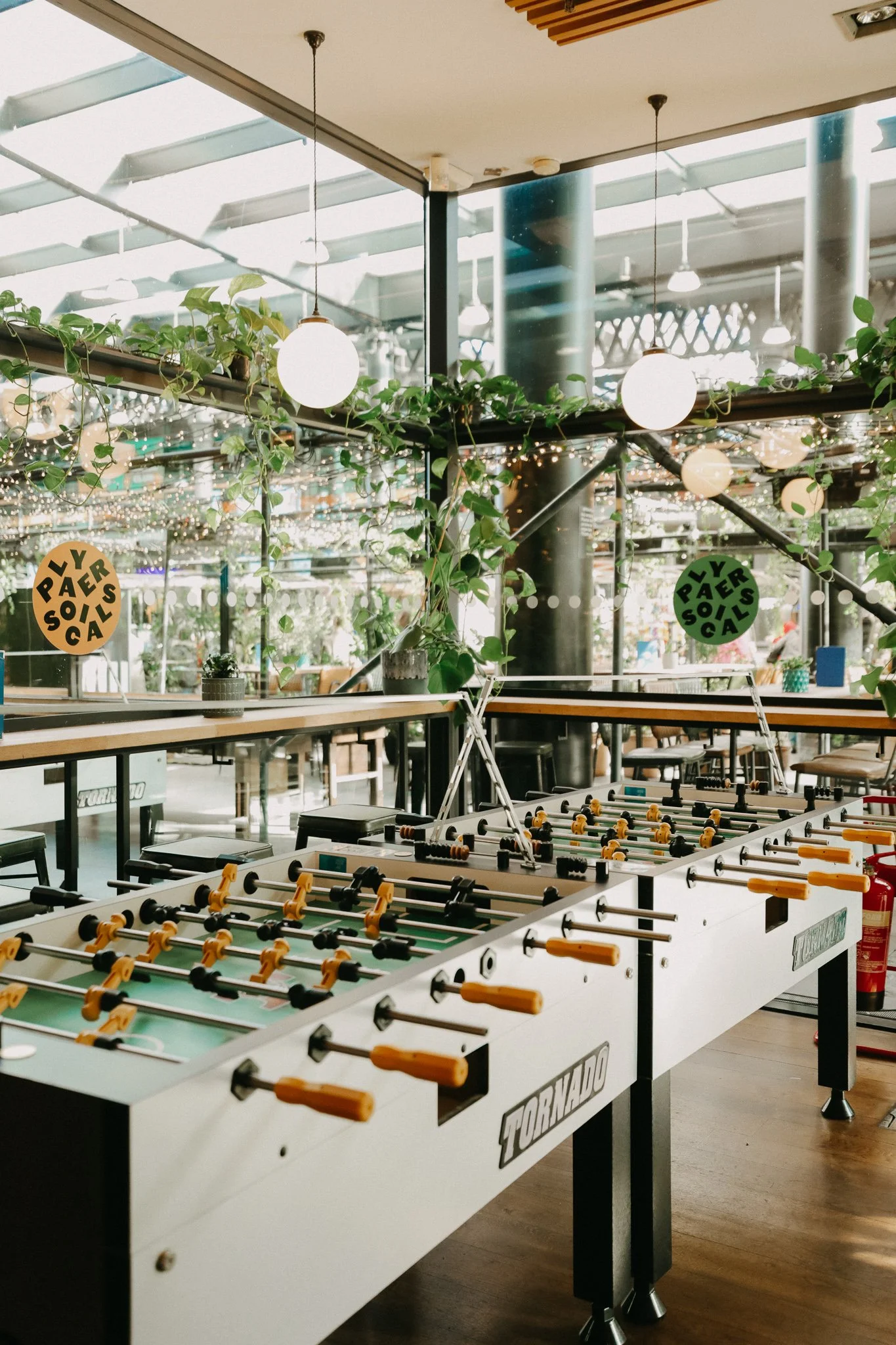 Indoor area with foosball tables, plants, hanging lights. Players Social Cocktail and Games Bar Spitalfields Market. Cocktail classes in London