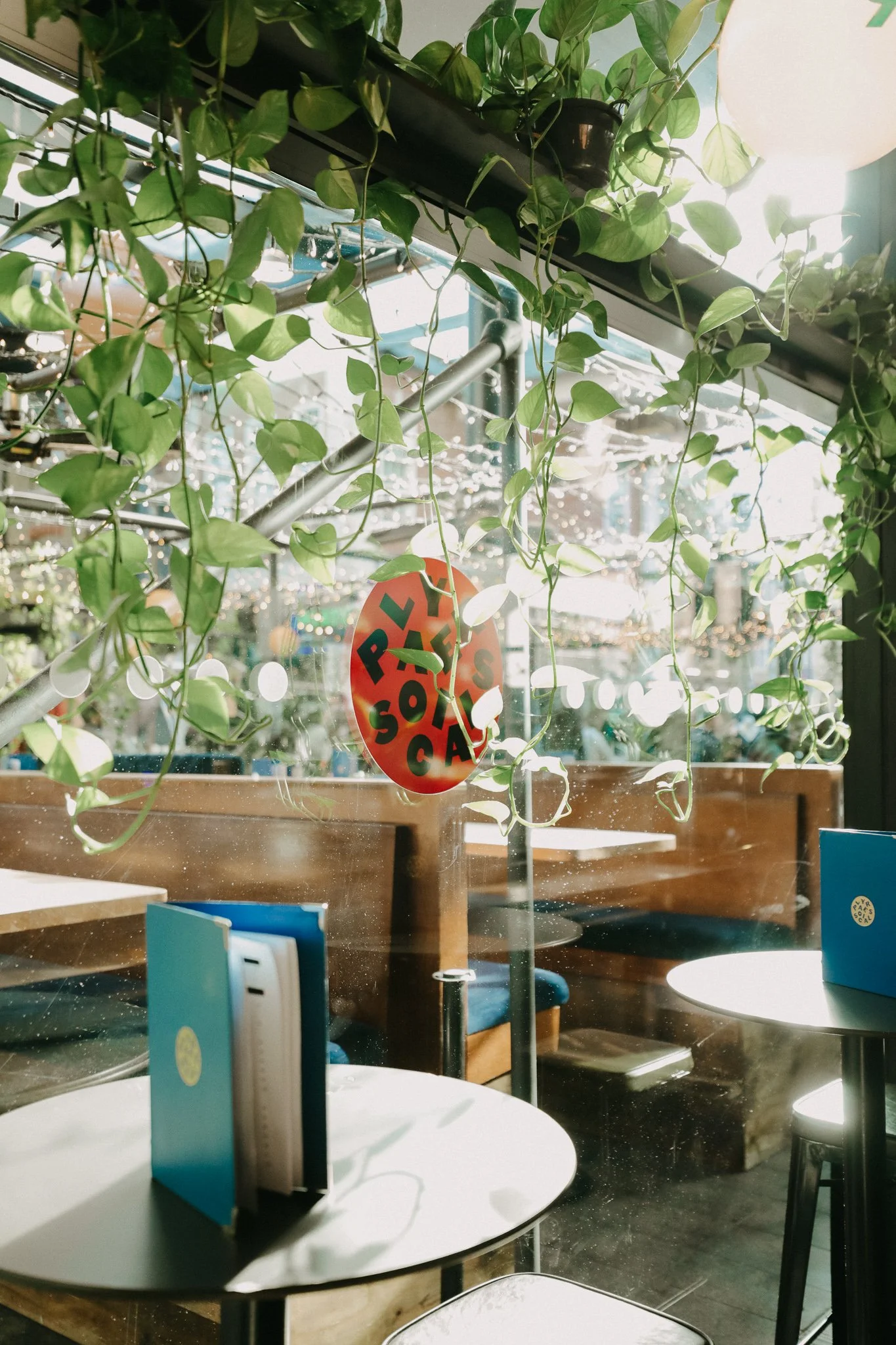 Interior of a bar with round white tables, blue menus, and lush green hanging plants. Natural light streams in through large windows. Players Social Cocktail and Games Bar Spitalfields Market. Cocktail classes in London