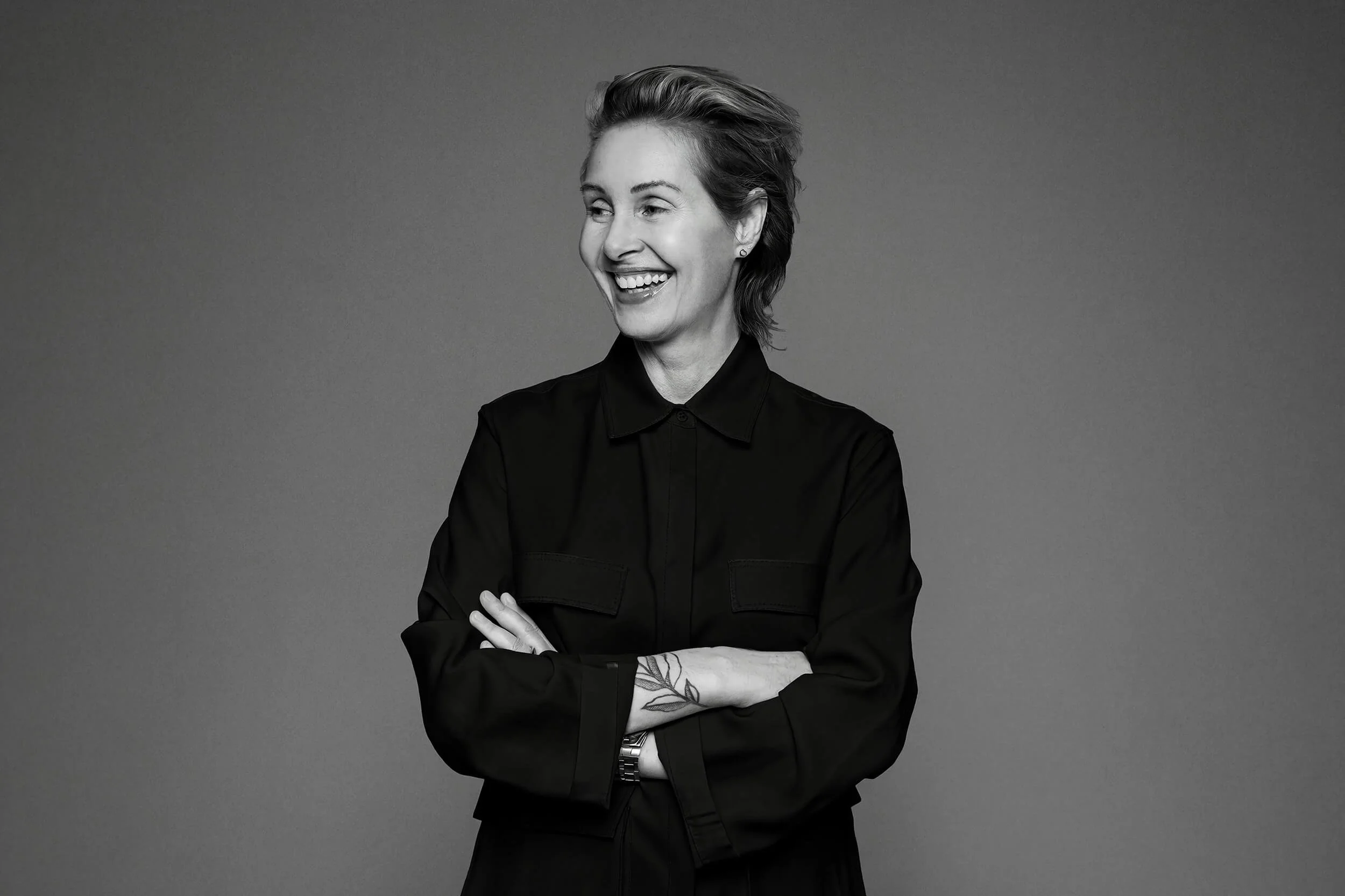 Black and white photo of a woman smiling with crossed arms, wearing a dark shirt, against a neutral background.