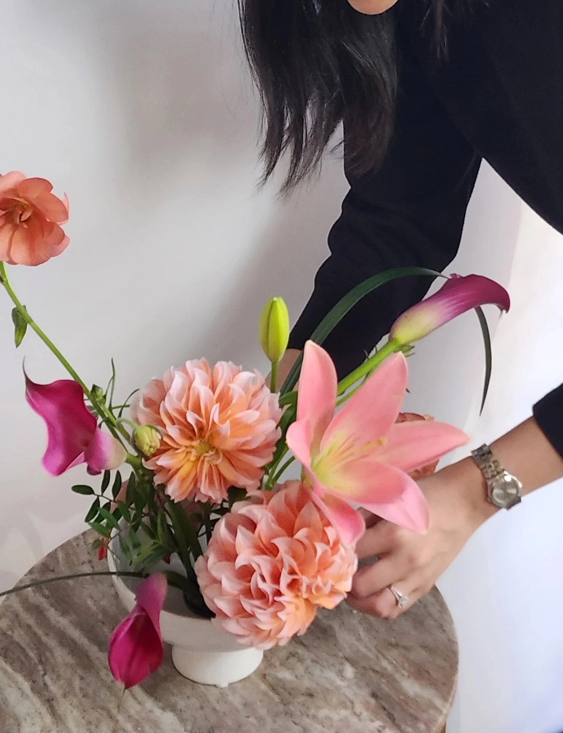 A woman with long dark hair in a black long sleeve shirt holding a vibrant bouquet of pink lilies, peach dahlias, and other flowers, placed on a beige stone table.