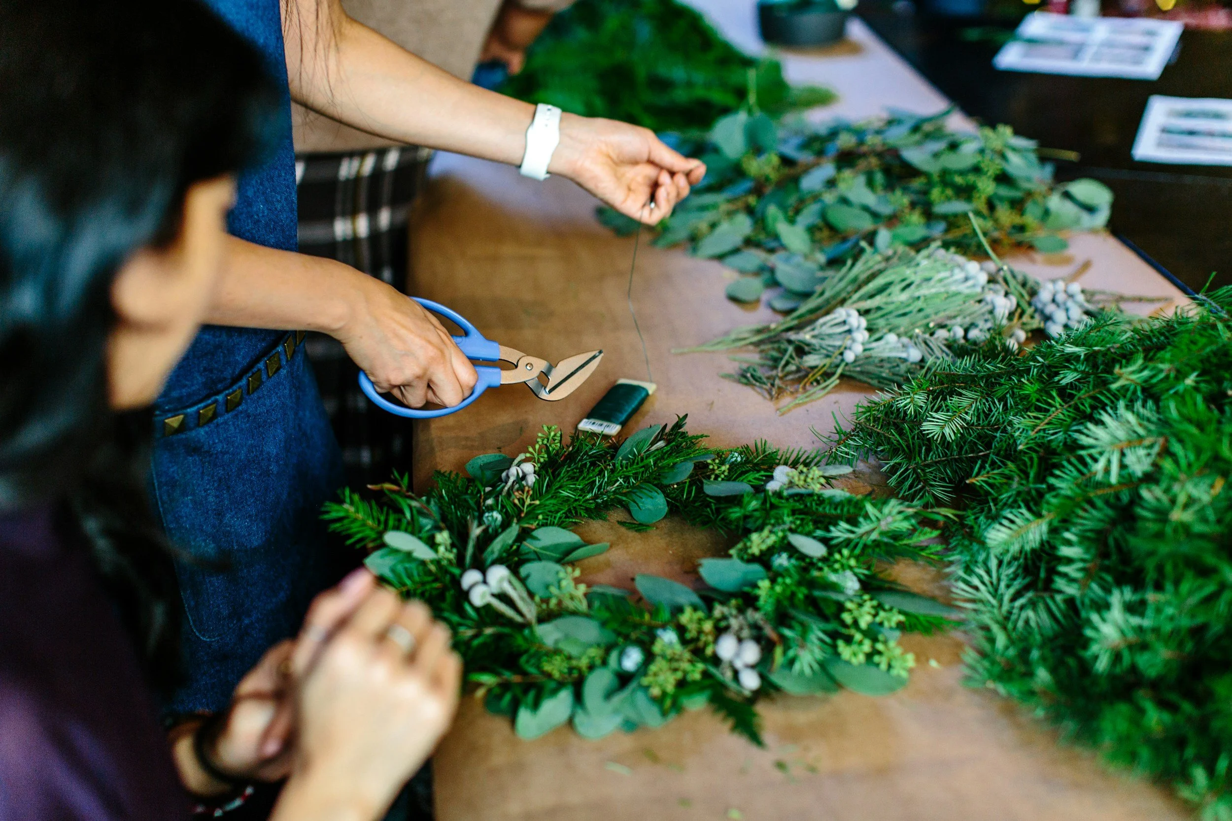 People working on a floral arrangement with various green foliage and white berries on a table.