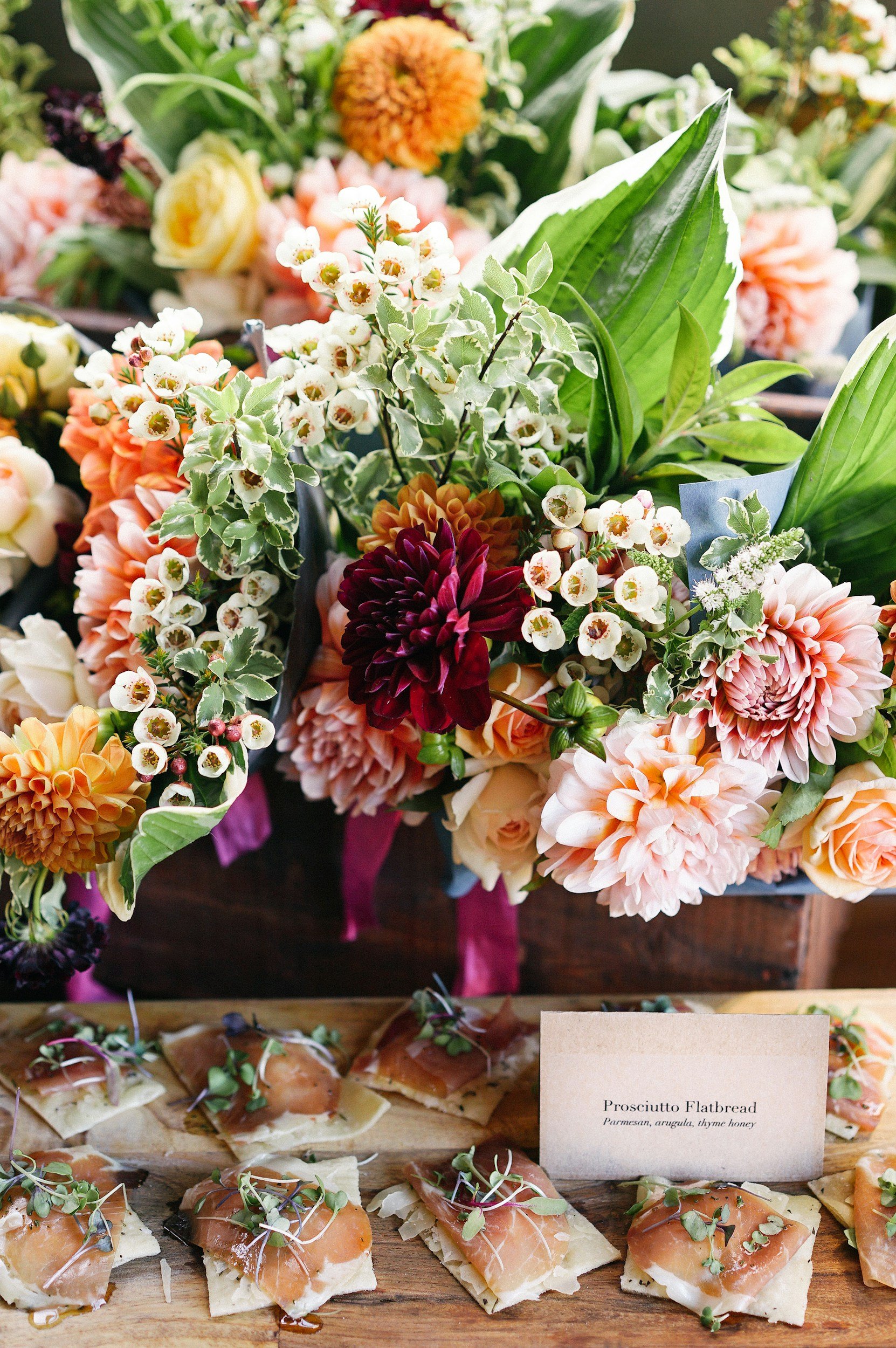 A display of assorted flowers including orange, pink, white, and purple blooms with green leaves, set above a wooden board with prosciutto flatbread topped with microgreens and cheese.