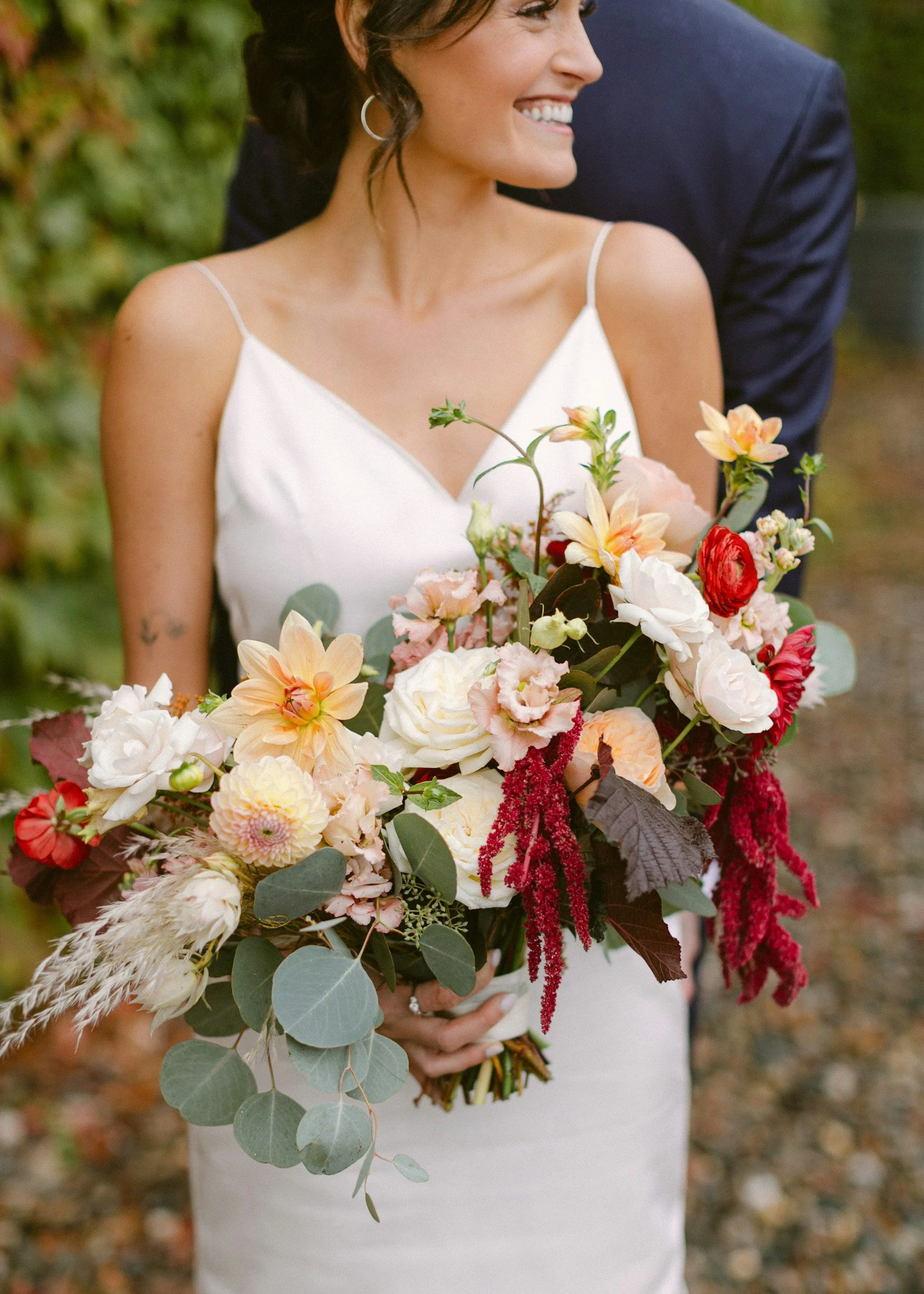 A smiling woman in a white dress holding a large bouquet of colorful flowers outdoors, with a man partially visible behind her.