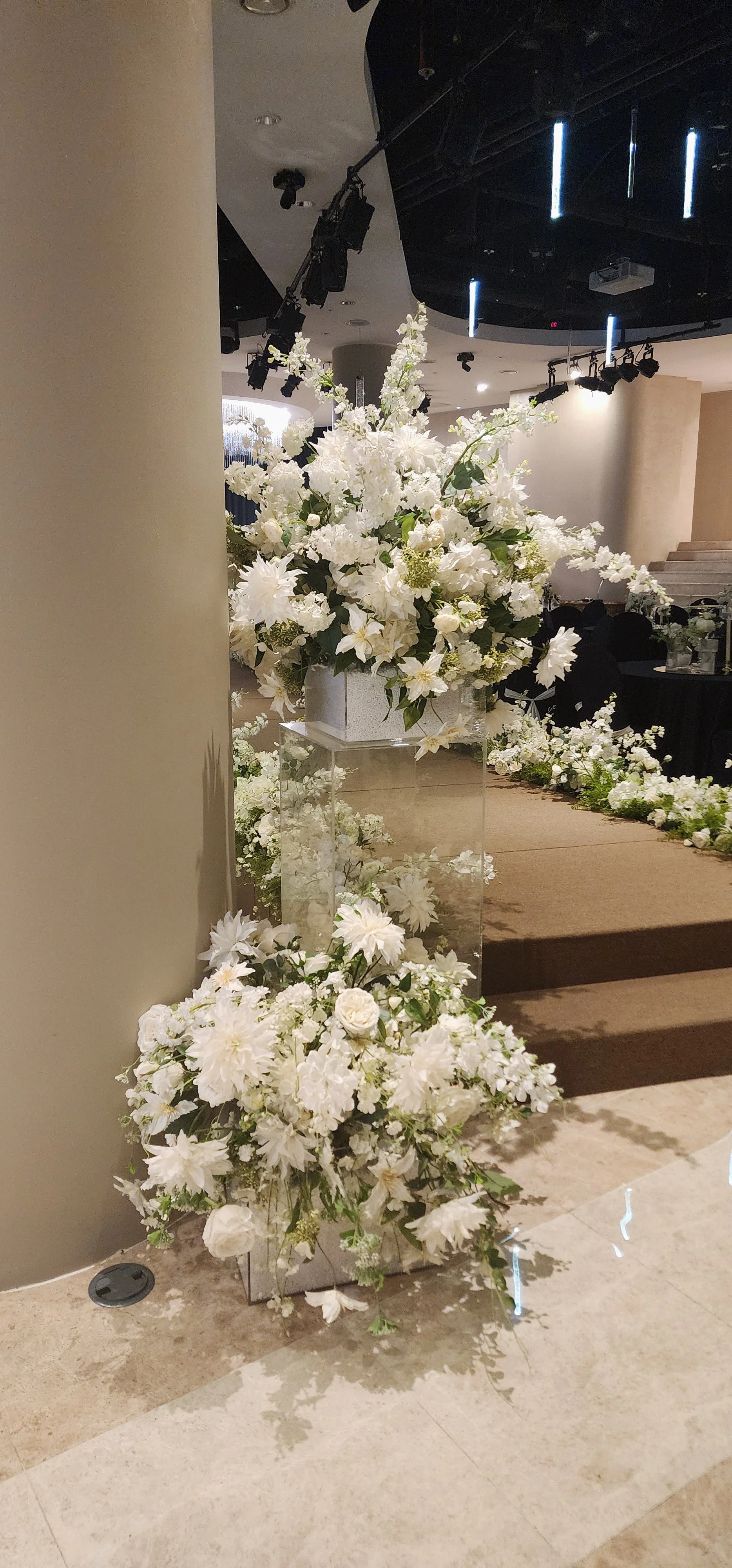A large, white floral arrangement on a glass stand in an event space, surrounded by additional white flowers and decorations, with a stage and ceiling lights in the background.