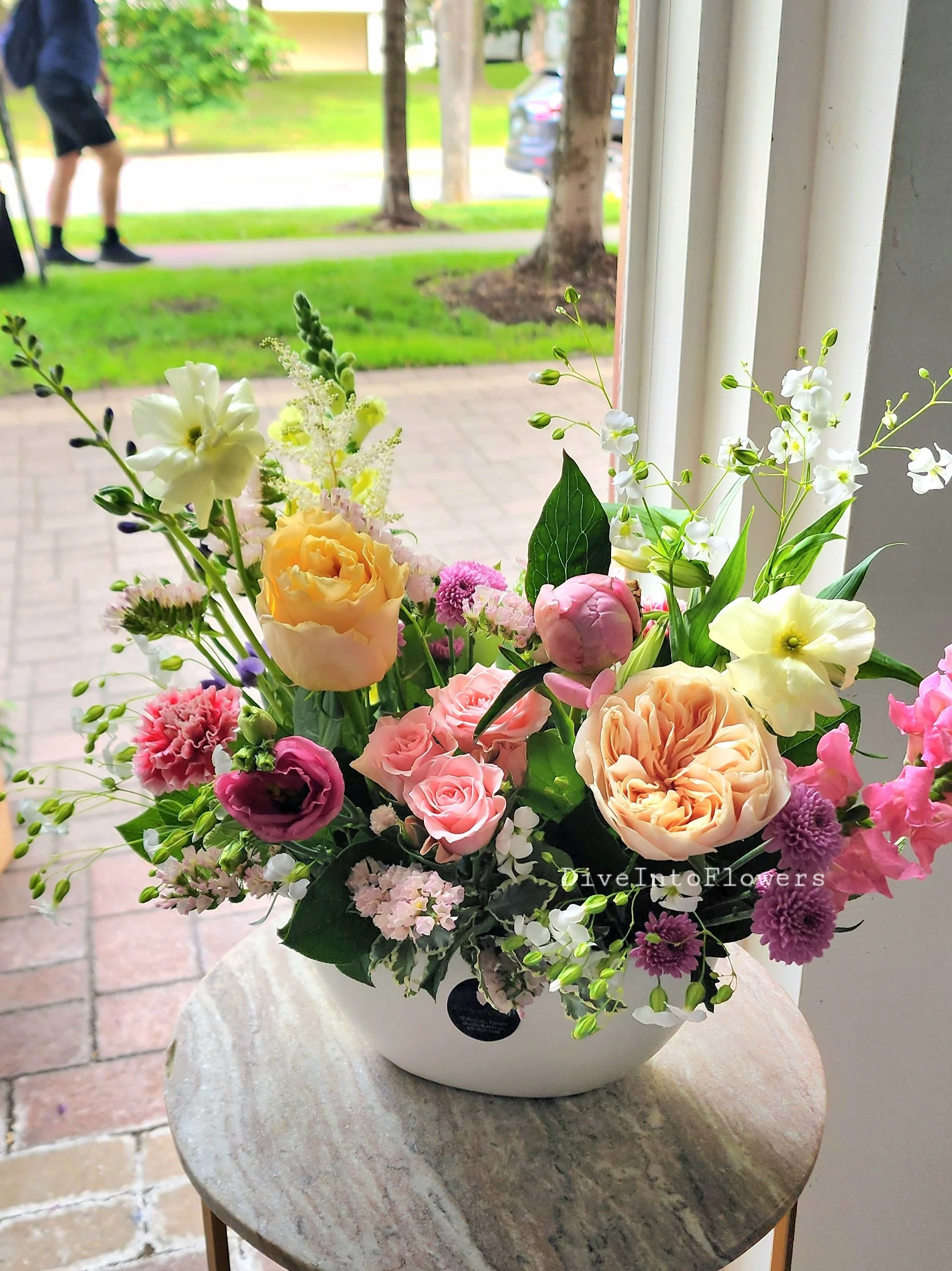 A bouquet of mixed flowers including roses, peonies, and various smaller blossoms, arranged in a white vase on a round stone table near a window. Outside, there are trees, a grassy area, and a person walking with a cane.