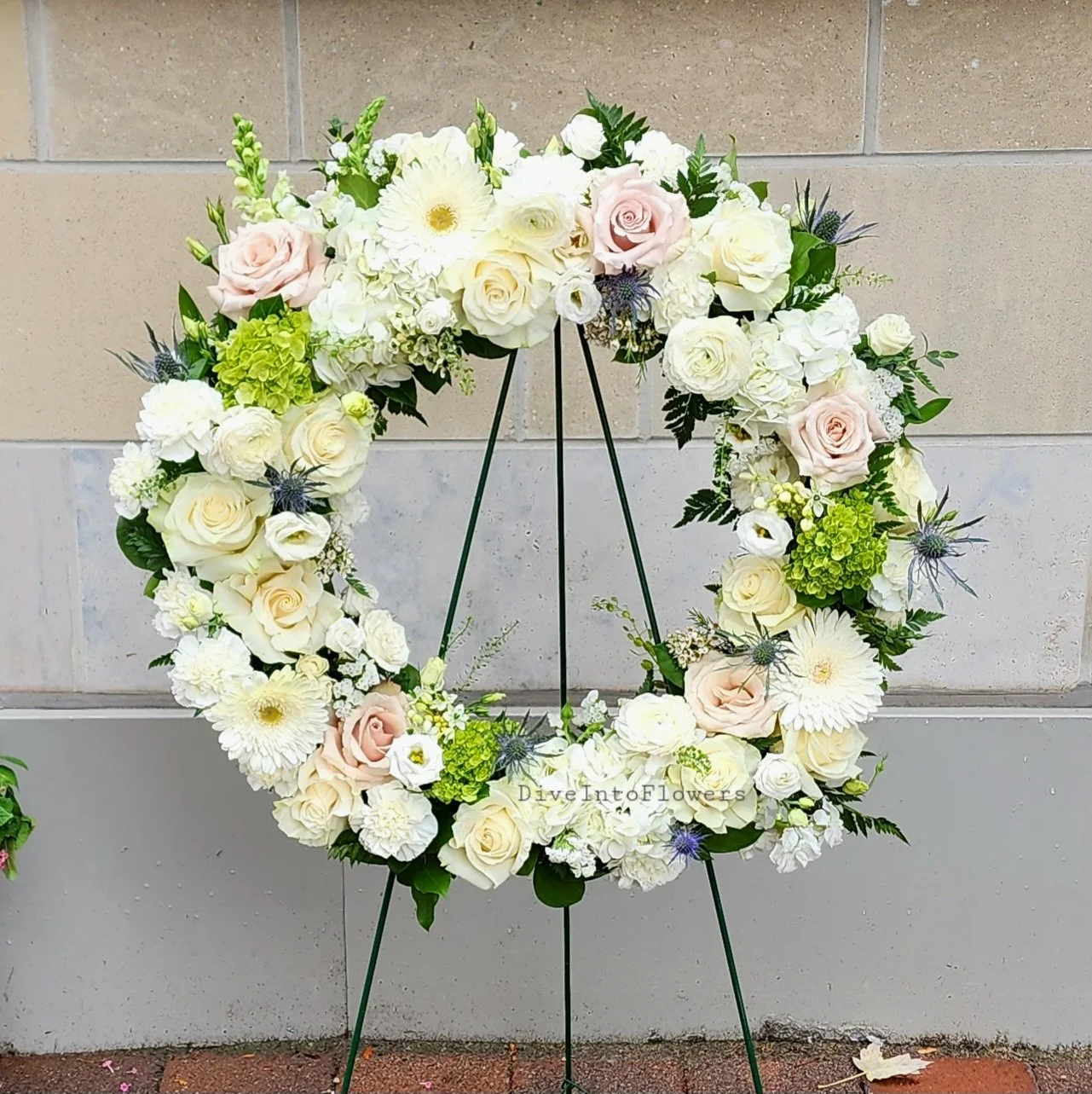 A wreath of white and light pink roses, white gerbera daisies, green hydrangeas, and various green foliage on a black metal stand against a beige brick wall.