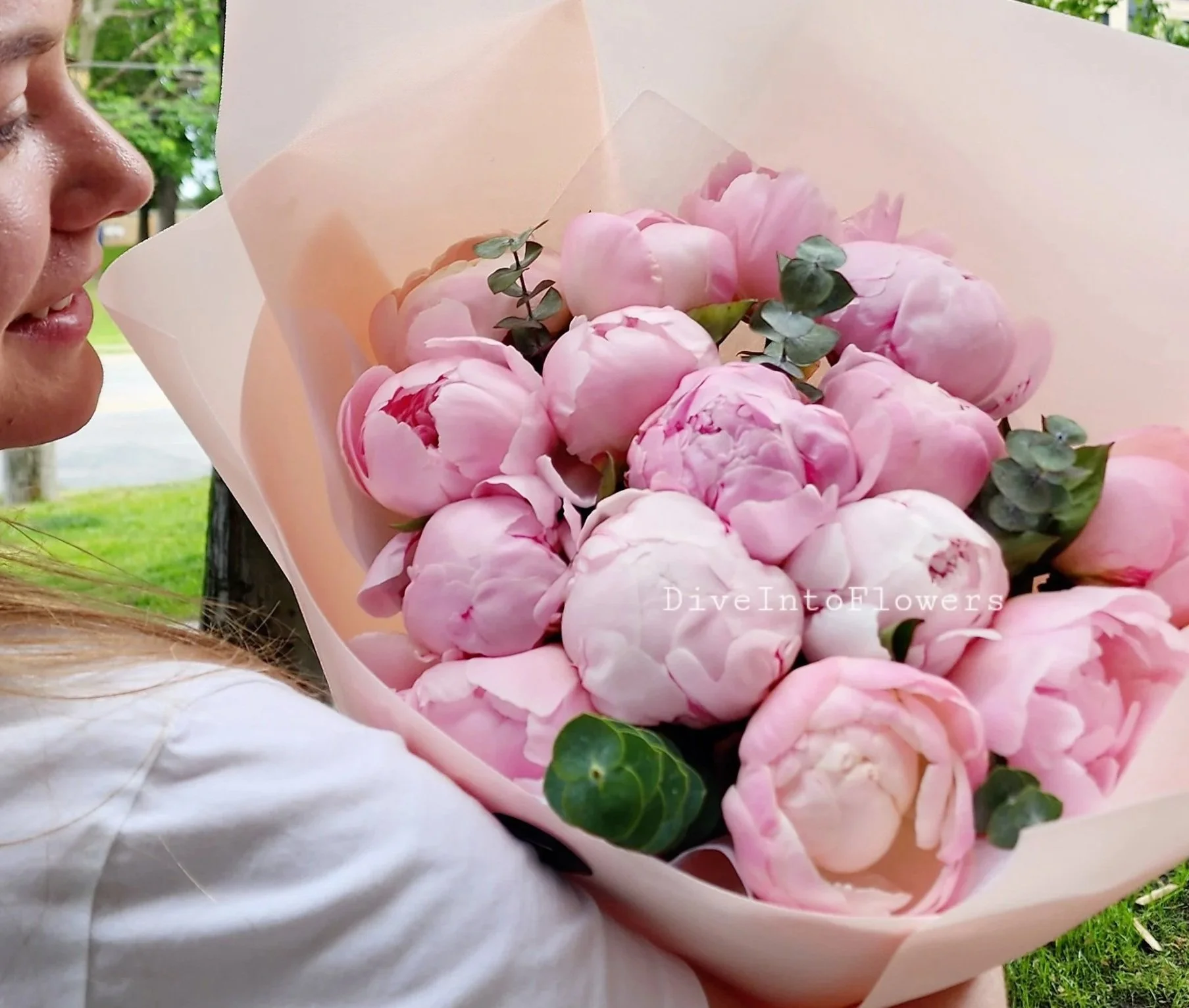 Close-up of a person holding a large bouquet of pink peonies wrapped in light pink paper, with greenery accents, outdoors with trees and grass in the background.