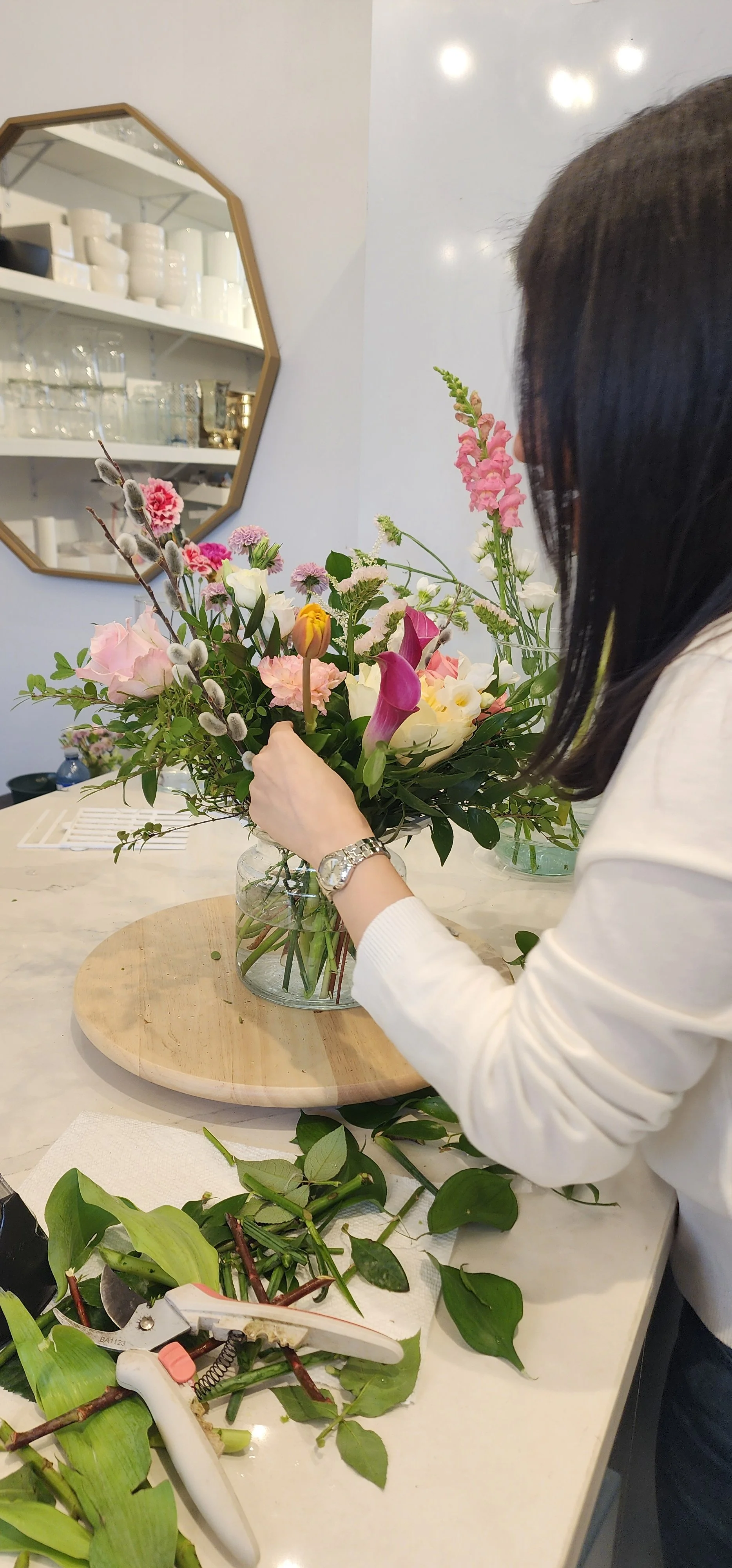 A woman arranging a bouquet of colorful flowers in a glass jar, with floral arranging tools and green leaves on the table, in a bright, modern room.